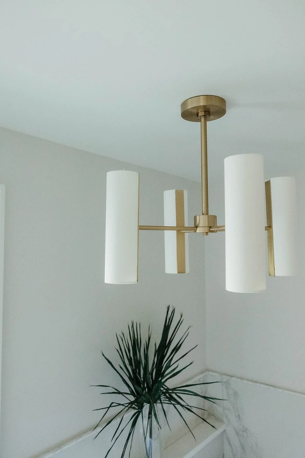 Close-up of a modern brass chandelier with three white cylindrical shades, hanging over the white marble wainscoting and a potted plant in the corner of the bathroom.