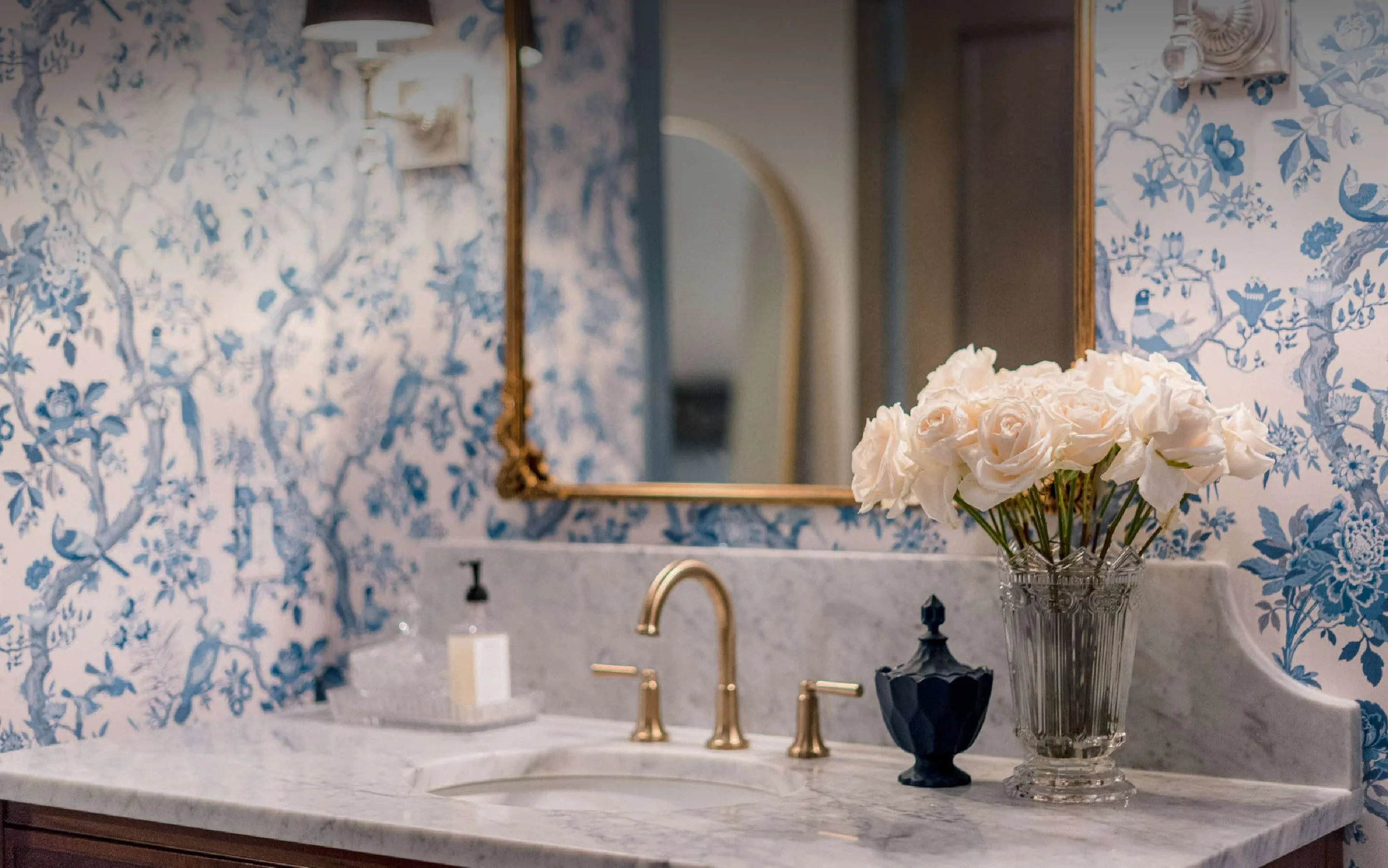 Traditional powder room with blue and white toile wallpaper, a marble countertop, and a brass widespread faucet. A vase of white roses sits next to a gold-framed mirror