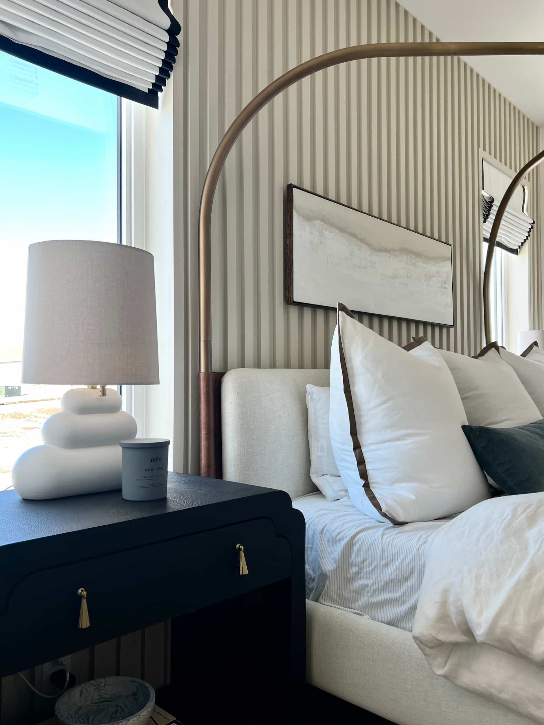 Close-up of the master bedroom nightstand with a black finish, featuring a unique stacked ceramic table lamp and a stack of towels. The slatted wood accent wall is visible behind the bed."