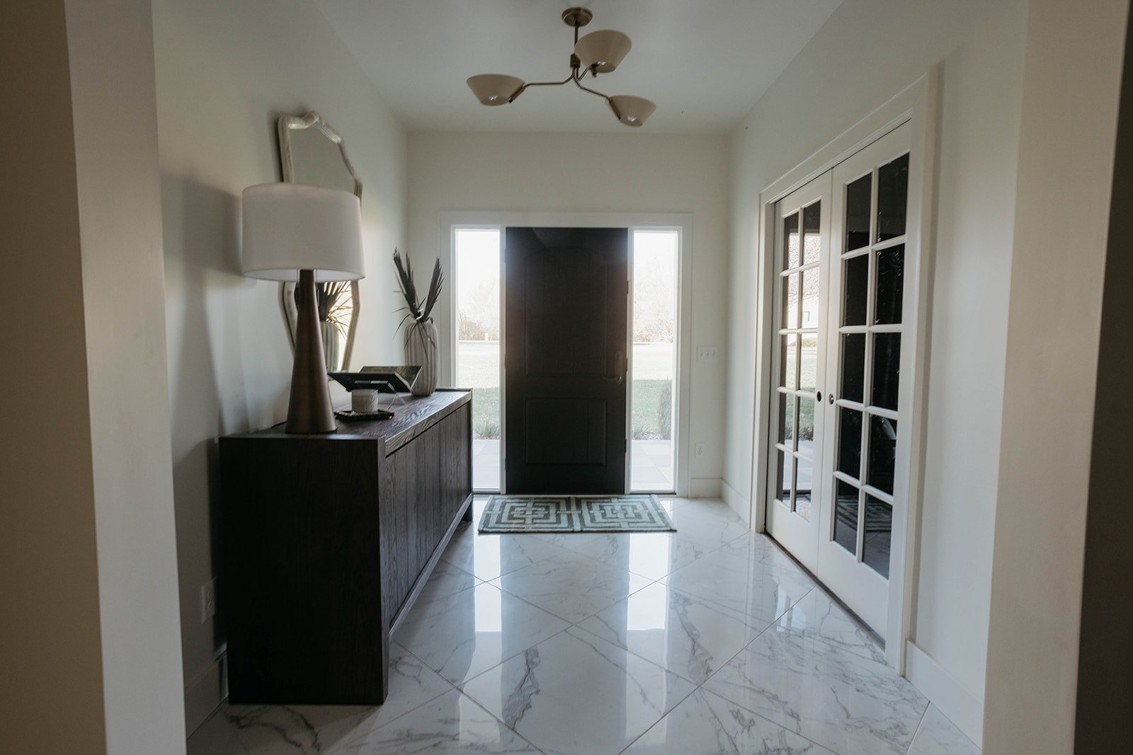 Modern entryway with marble tile floor, featuring a long dark wood console cabinet styled with a mirror, lamp, and vase. The front door and French doors are visible.
