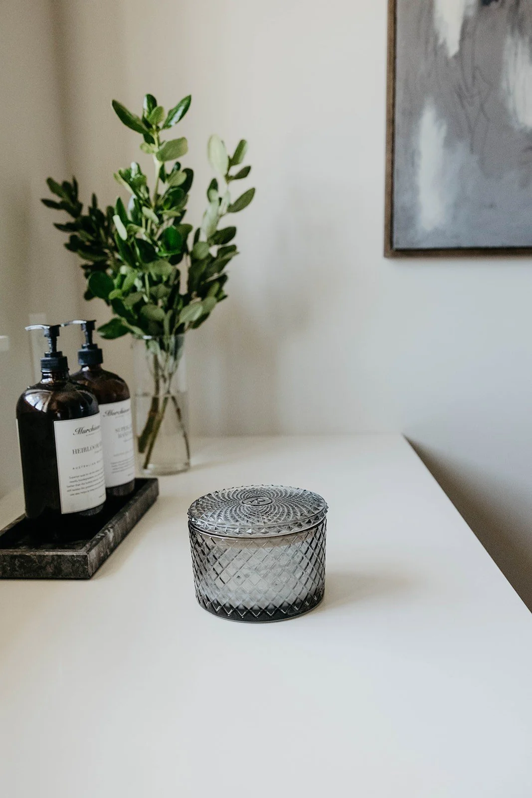 Close-up of the clean white laundry room countertop styled with a black tray of soap dispensers, a vase of greenery, and a textured glass storage jar.