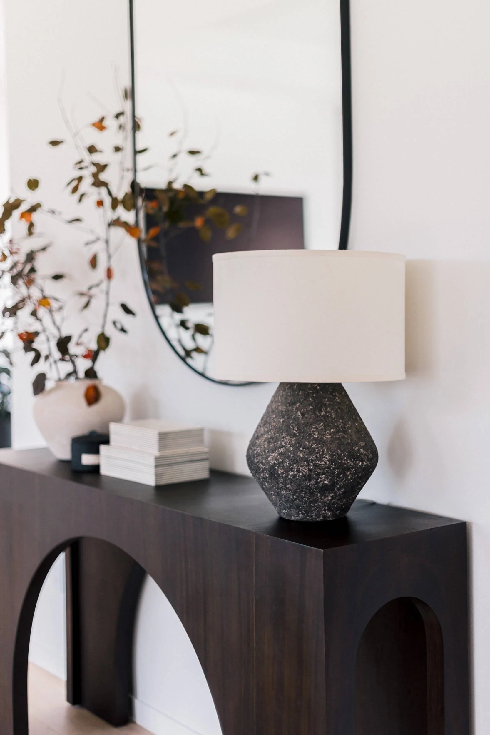Close-up of the dark wood entryway console table, showing the speckled black lamp, stack of white books, and the reflection in the black-framed mirror.
