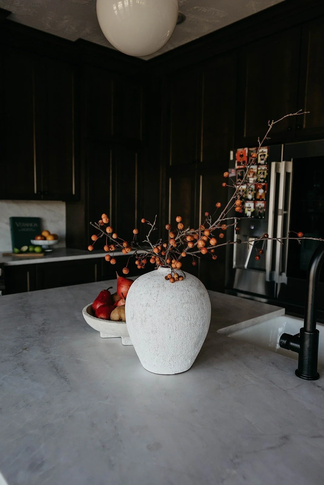 View of the kitchen countertop styled with a textured white vase holding cranberry branches and a bowl of fruit, with the dark cabinetry in the background.