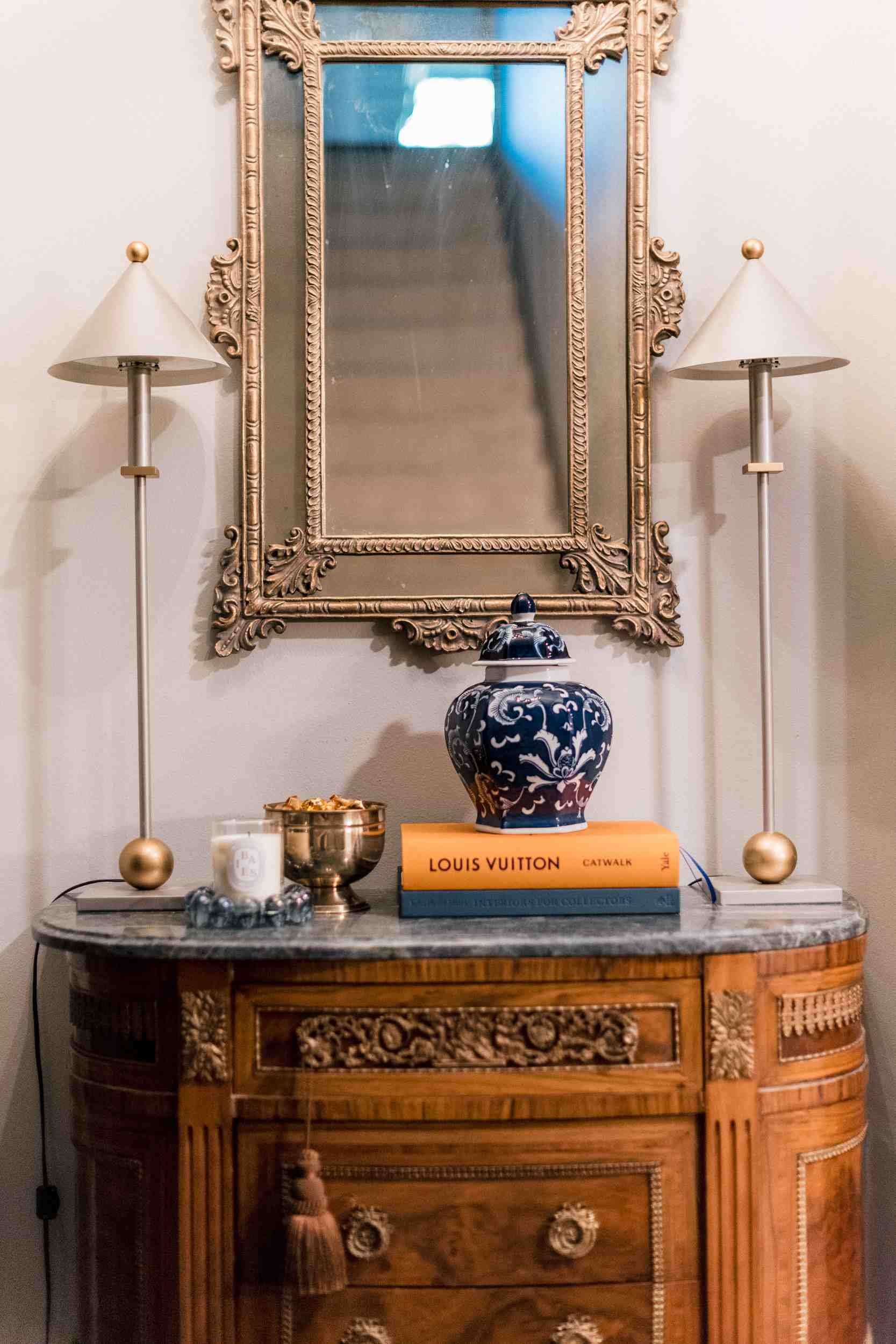 Traditional entryway featuring a carved wood antique marble-top console, styled with a gilt mirror, two tall lamps, and books.