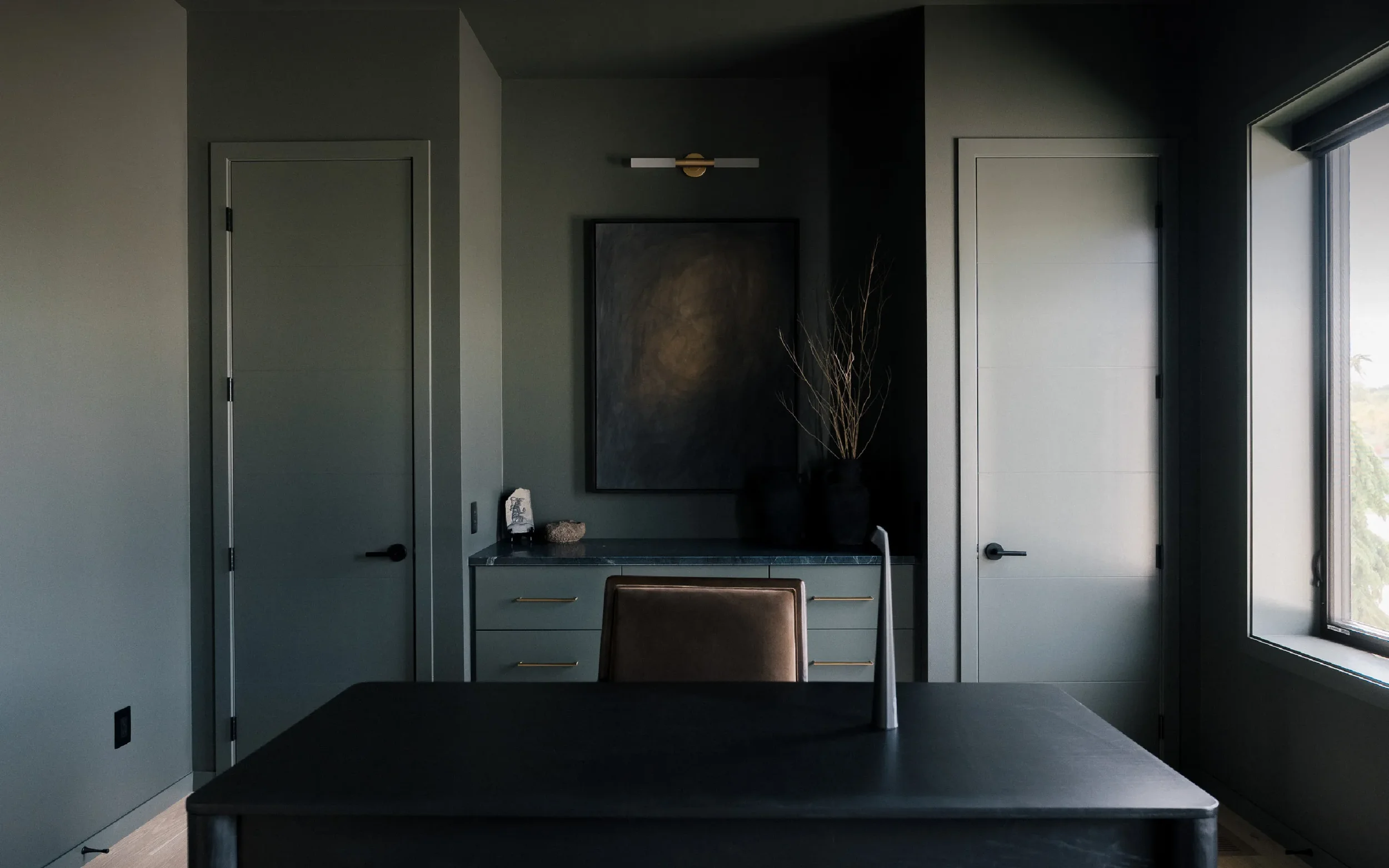 Modern office with dark walls, a table with a brown chair, a side cabinet with decorative items, and a large window.