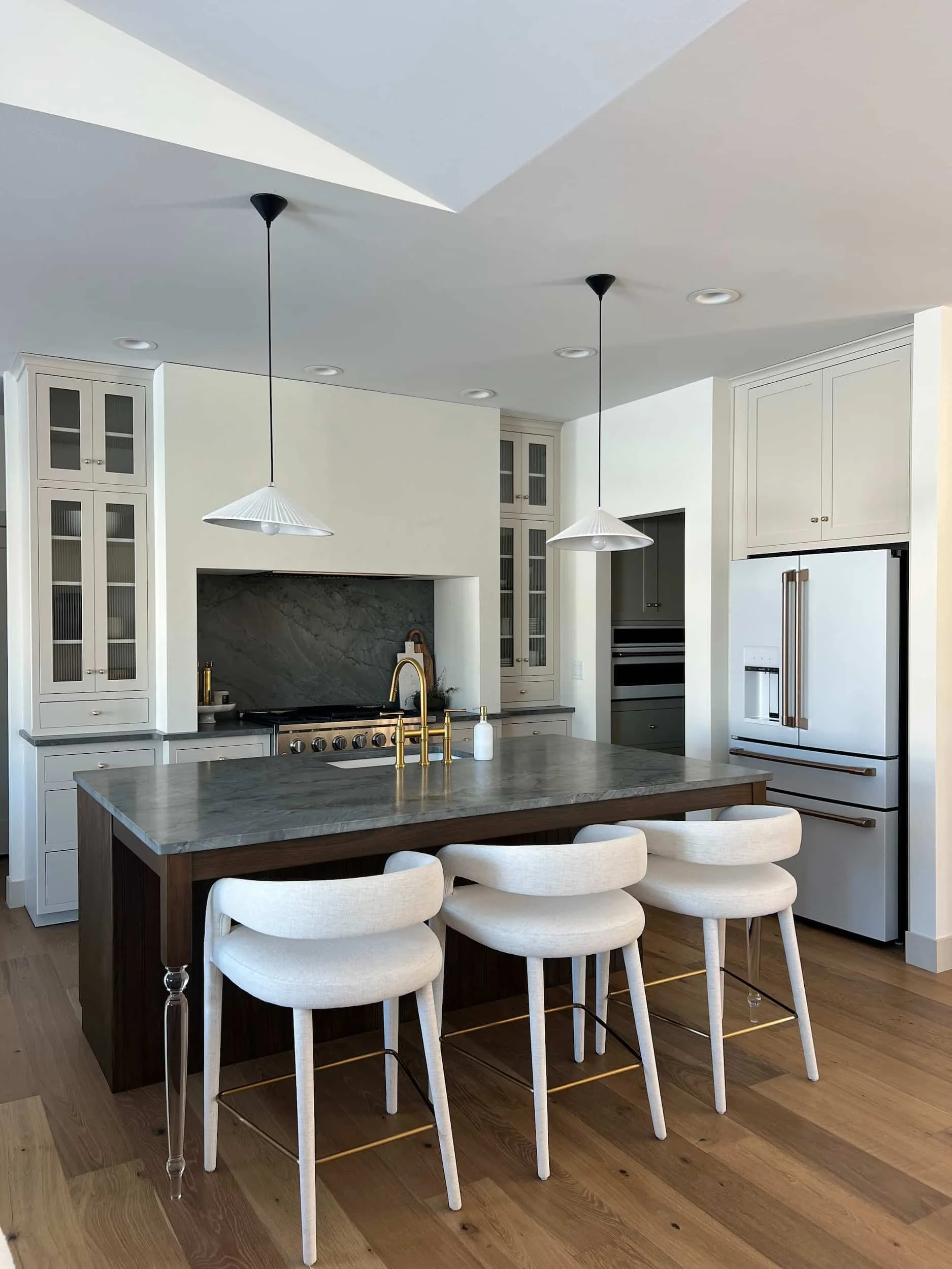 Spacious modern kitchen featuring white shaker cabinetry, a dark wood waterfall island, white marble backsplash, and three white curved counter stools.