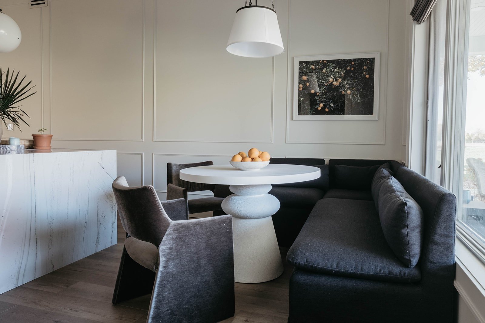 Close-up of the white round dining table in the nook, styled with a white bowl of oranges beneath a framed citrus artwork.