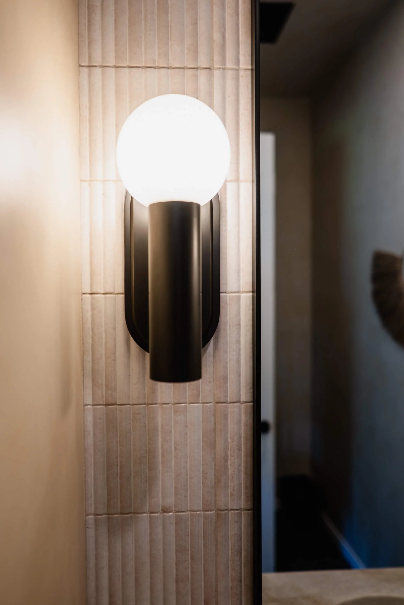 Close-up of a modern matte black wall sconce with a globe light, set against vertical light beige tiled walls.