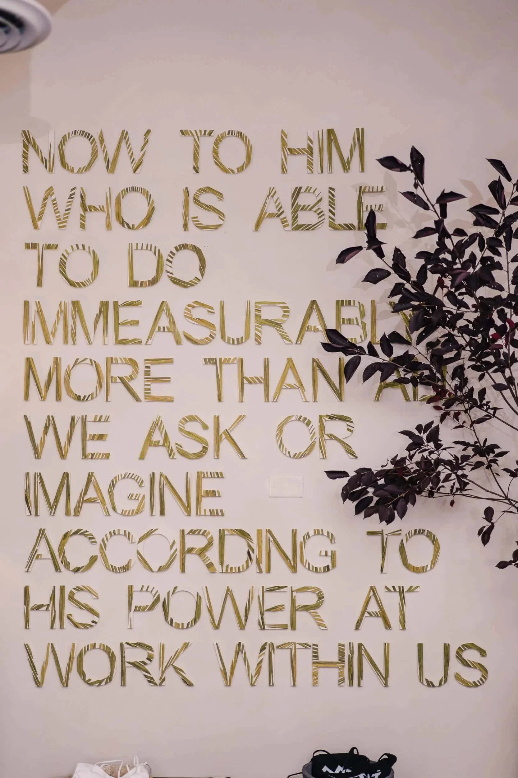 Motivational wall art with a biblical quote, 'Now to him who is able to do immeasurably more than all we ask or imagine according to his power that is at work within us,' rendered in gold 3D lettering on a light wall.