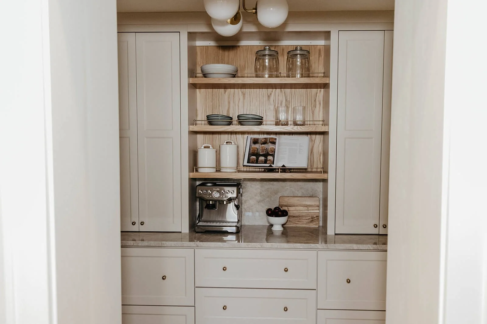 View of a neat, organized walk-in pantry or large utility cabinet with light gray or beige cabinetry and open wood shelving displaying serving ware and a coffee machine.