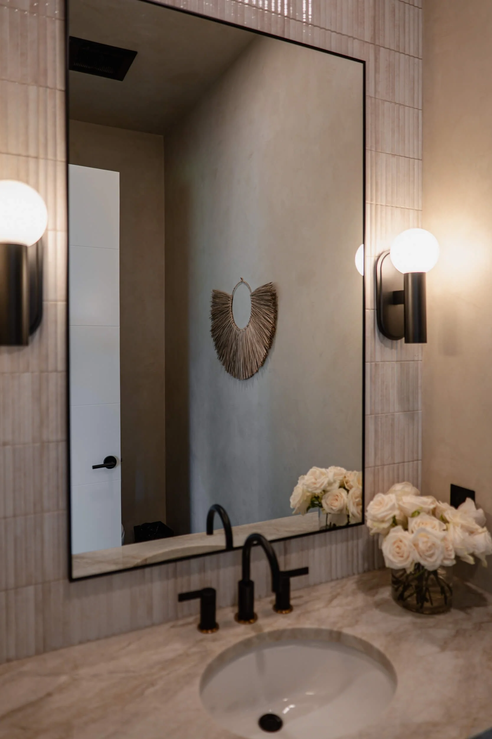Contemporary bathroom vanity with a light beige marble countertop, matte black faucet, large black-framed mirror, and a decorative wicker wall hanging.
