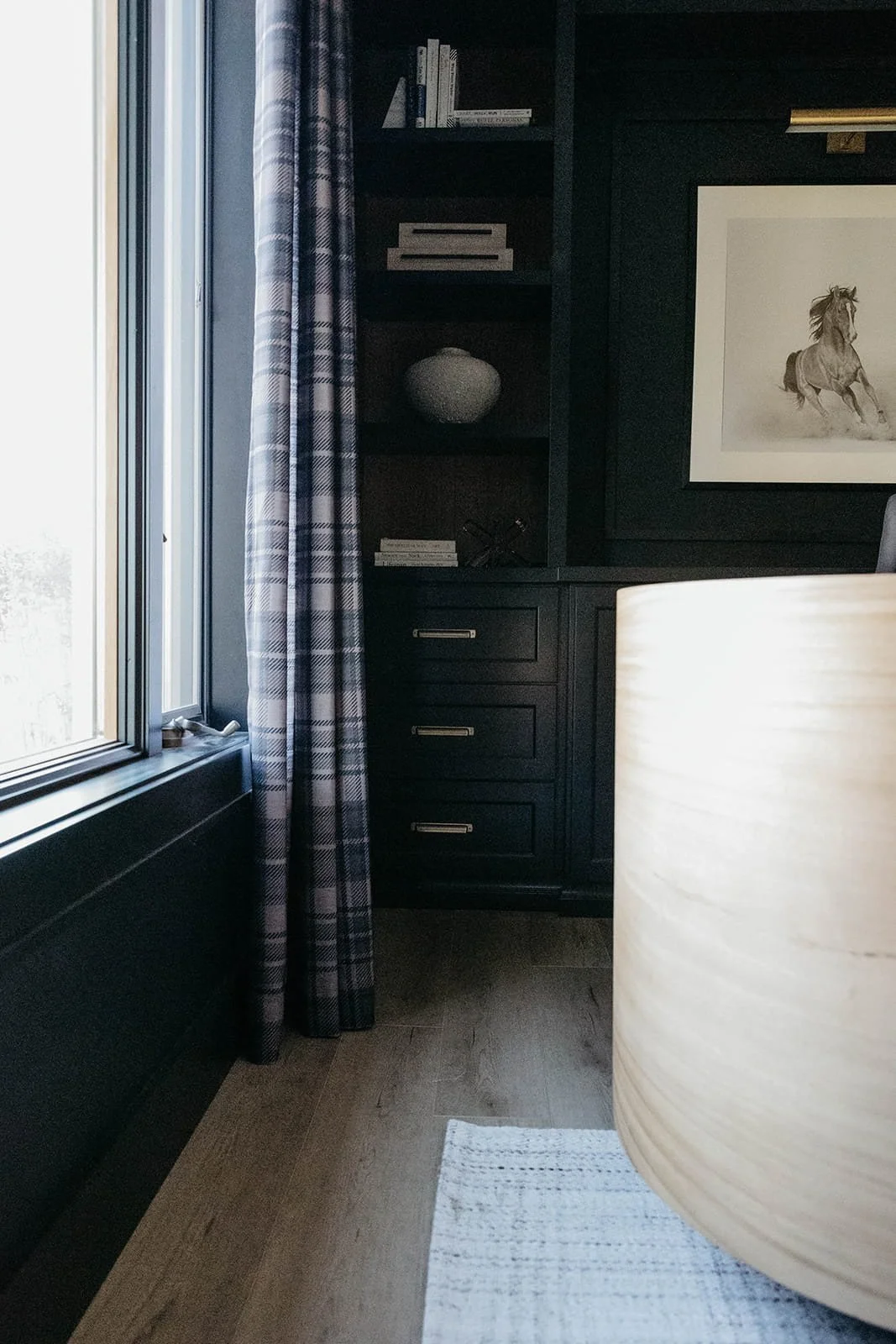 View of the dark built-in office cabinetry and window with a plaid curtain. A light wood curved desk is visible on the right, providing contrast to the dark walls.