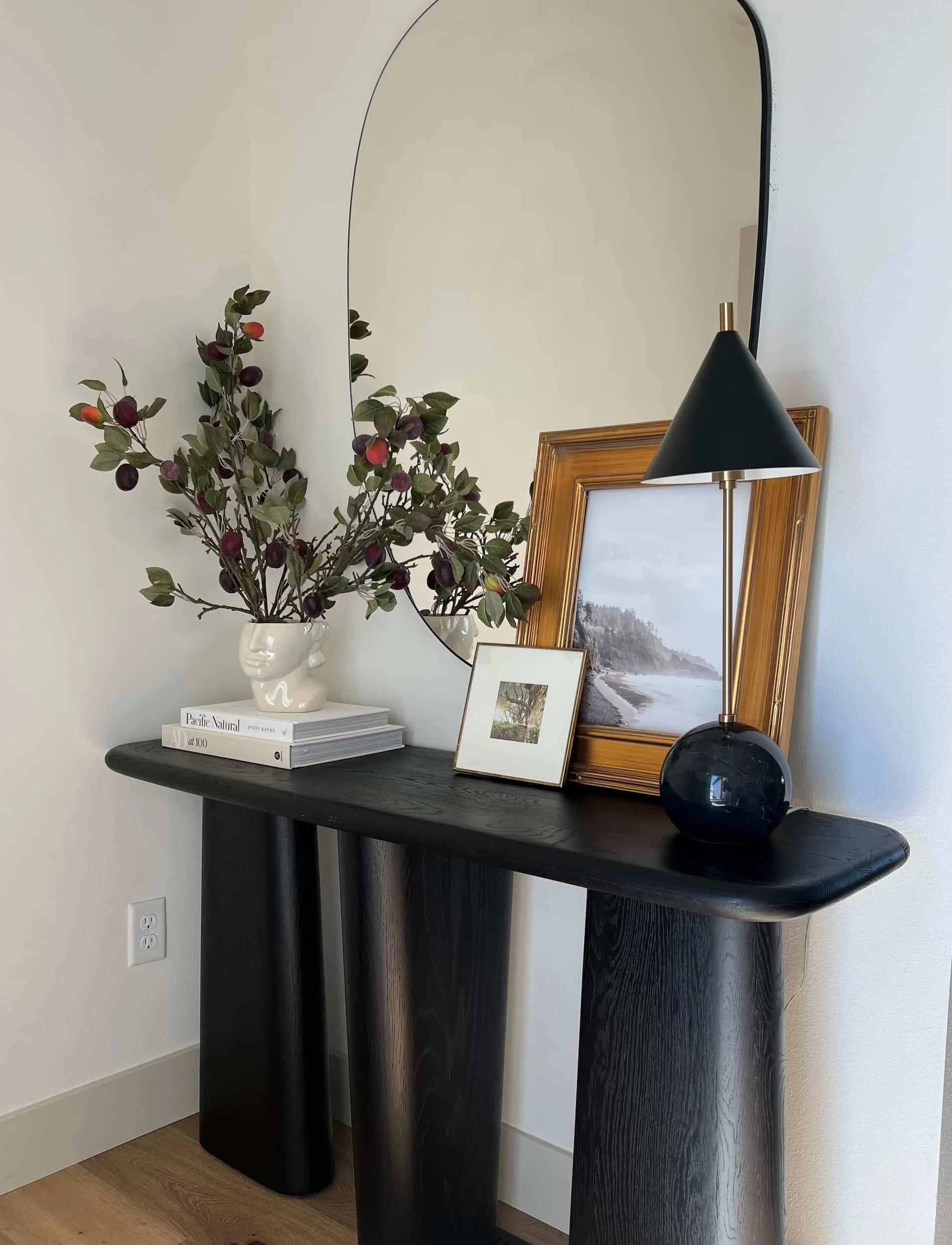 Close-up of a modern console table with a sculptural dark wood curved base, styled with a black and gold lamp, a vase of berries, and framed artwork.