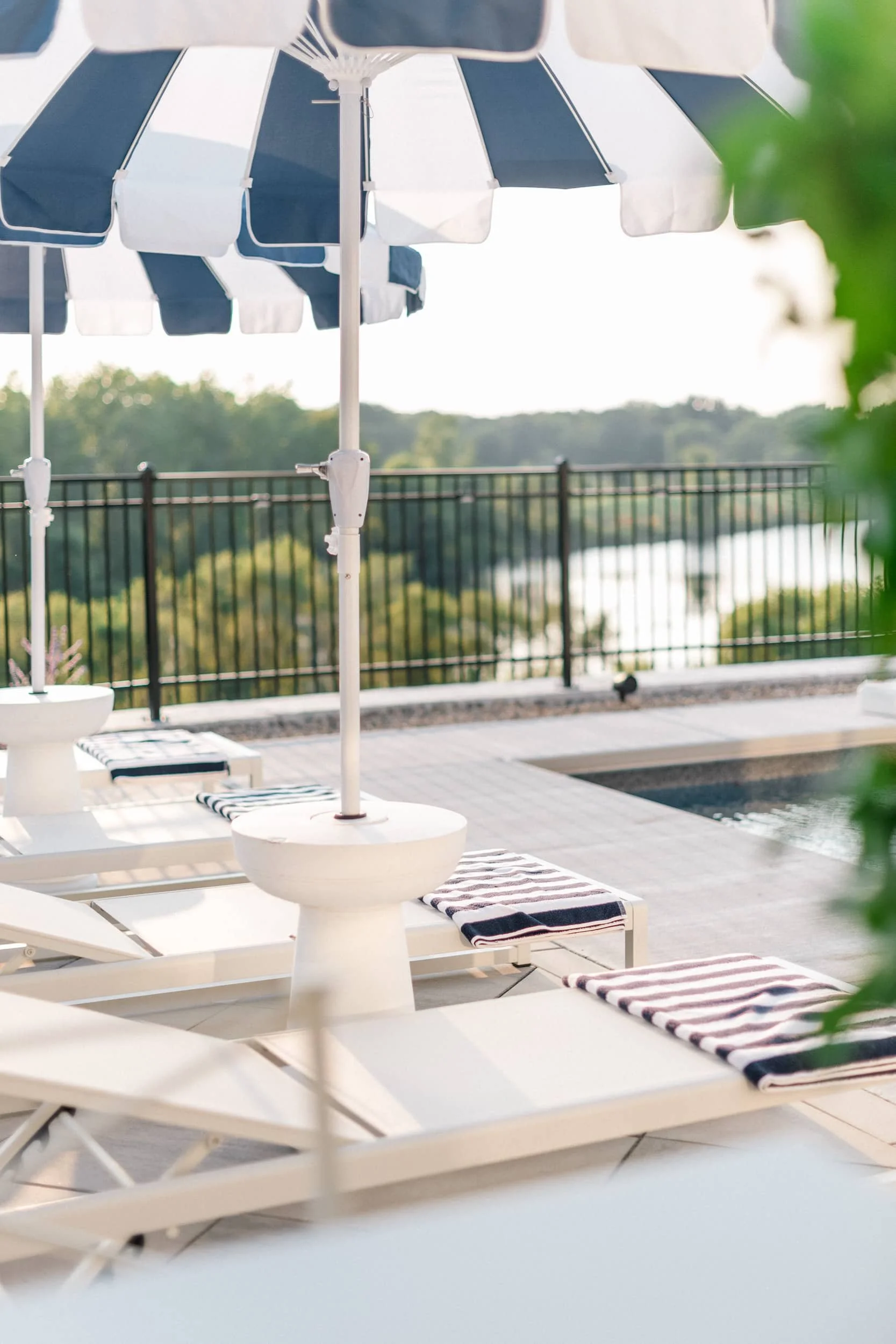 Close-up of white chaise lounges with striped cushions and a side table, under a partially shaded navy and white umbrella on the poolside patio.