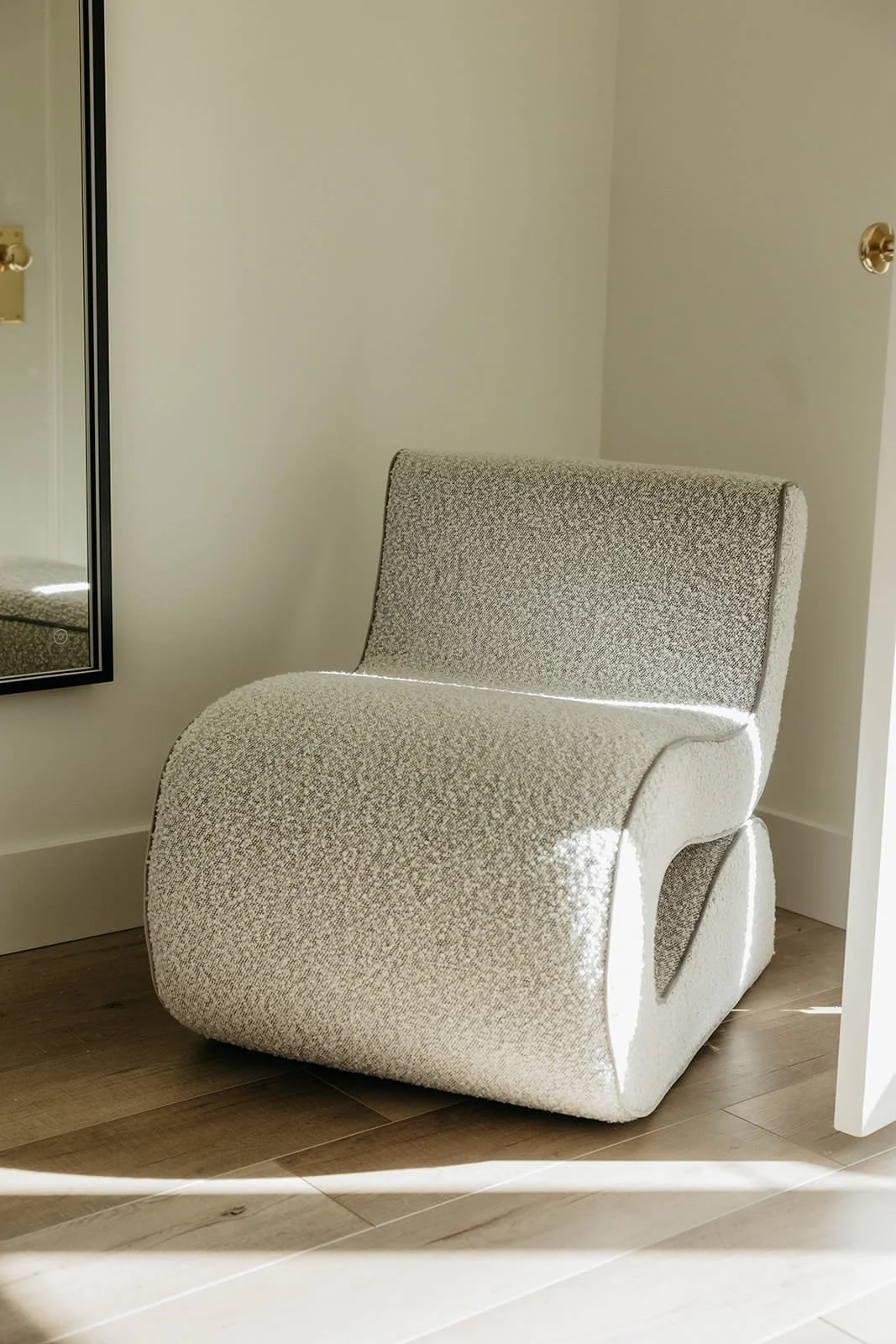 Close-up of a sculptural accent chair upholstered in white bouclé fabric, sitting on light wood flooring in a sunny corner.