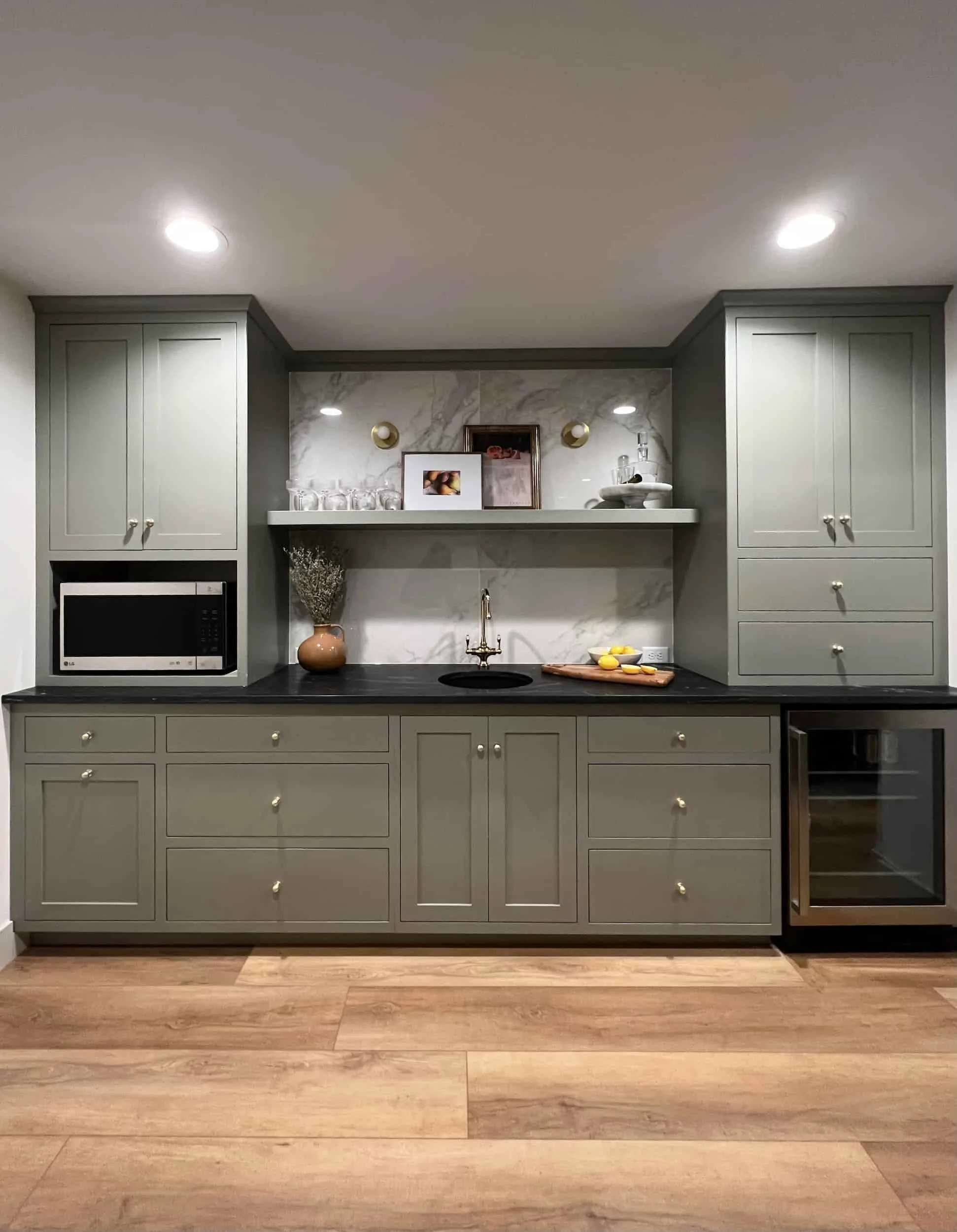 Stylish wet bar area with sage green cabinetry, a dark countertop, an integrated beverage fridge, and a marble slab backsplash with floating wood shelves.
