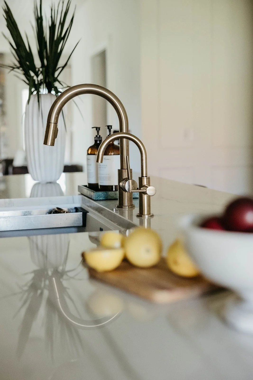 Tight close-up of a brushed gold kitchen faucet and prep faucet reflecting on the countertop, next to sliced lemons and a white vase with greenery.