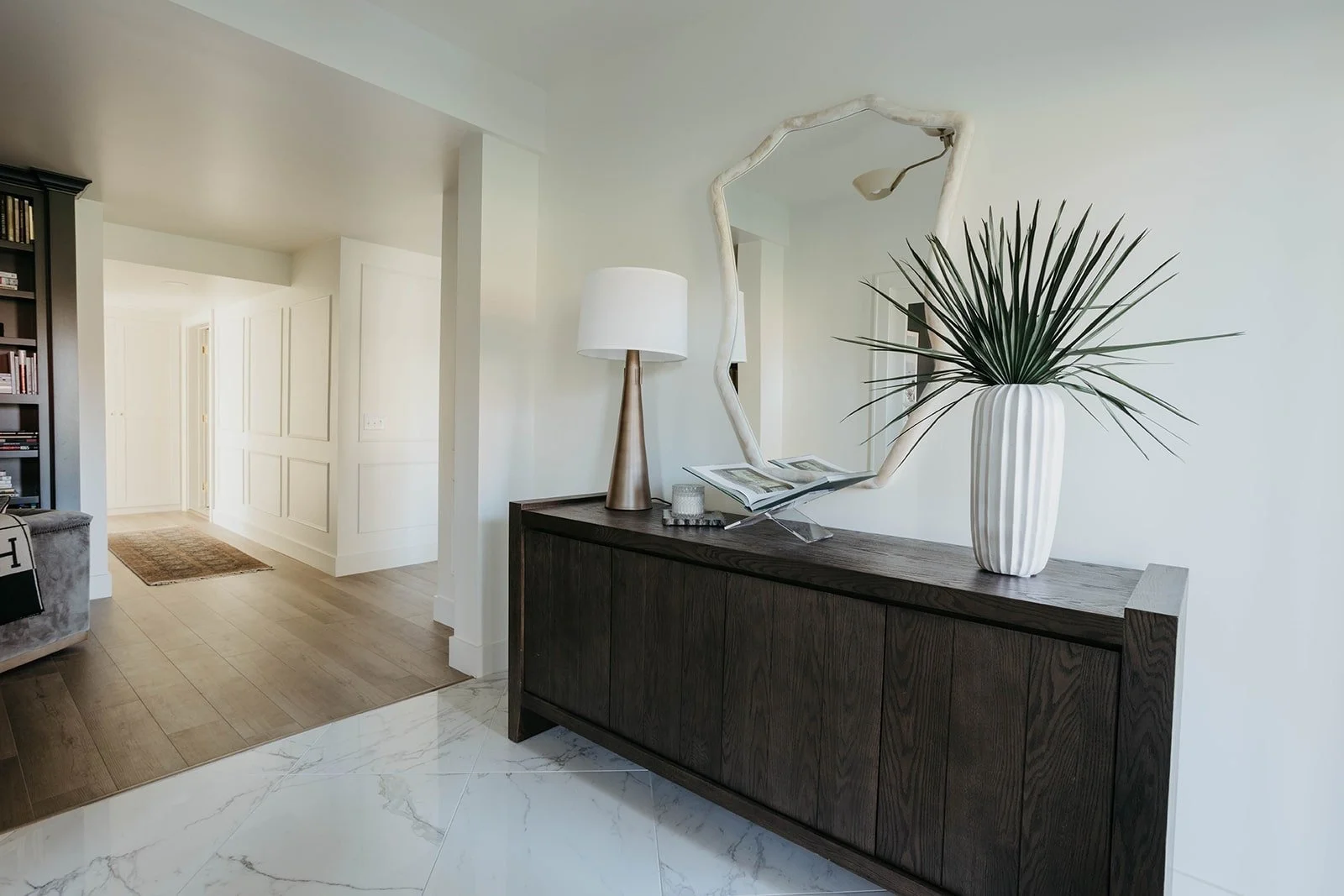 Entryway perspective showing the dark wood console and mirror, with a contrast between the white marble tile and hardwood flooring in the adjacent rooms.