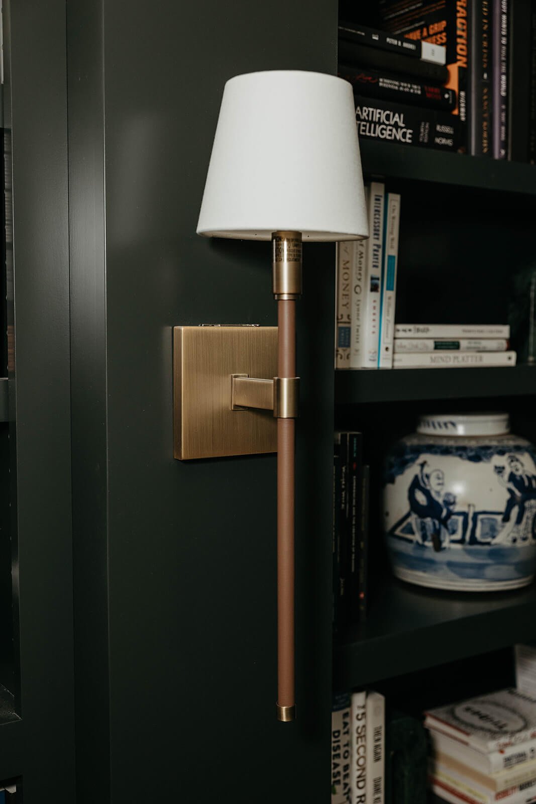 Close-up of a brass and white wall sconce mounted on the dark shelving, illuminating a stack of books and a blue and white chinoiserie vase.