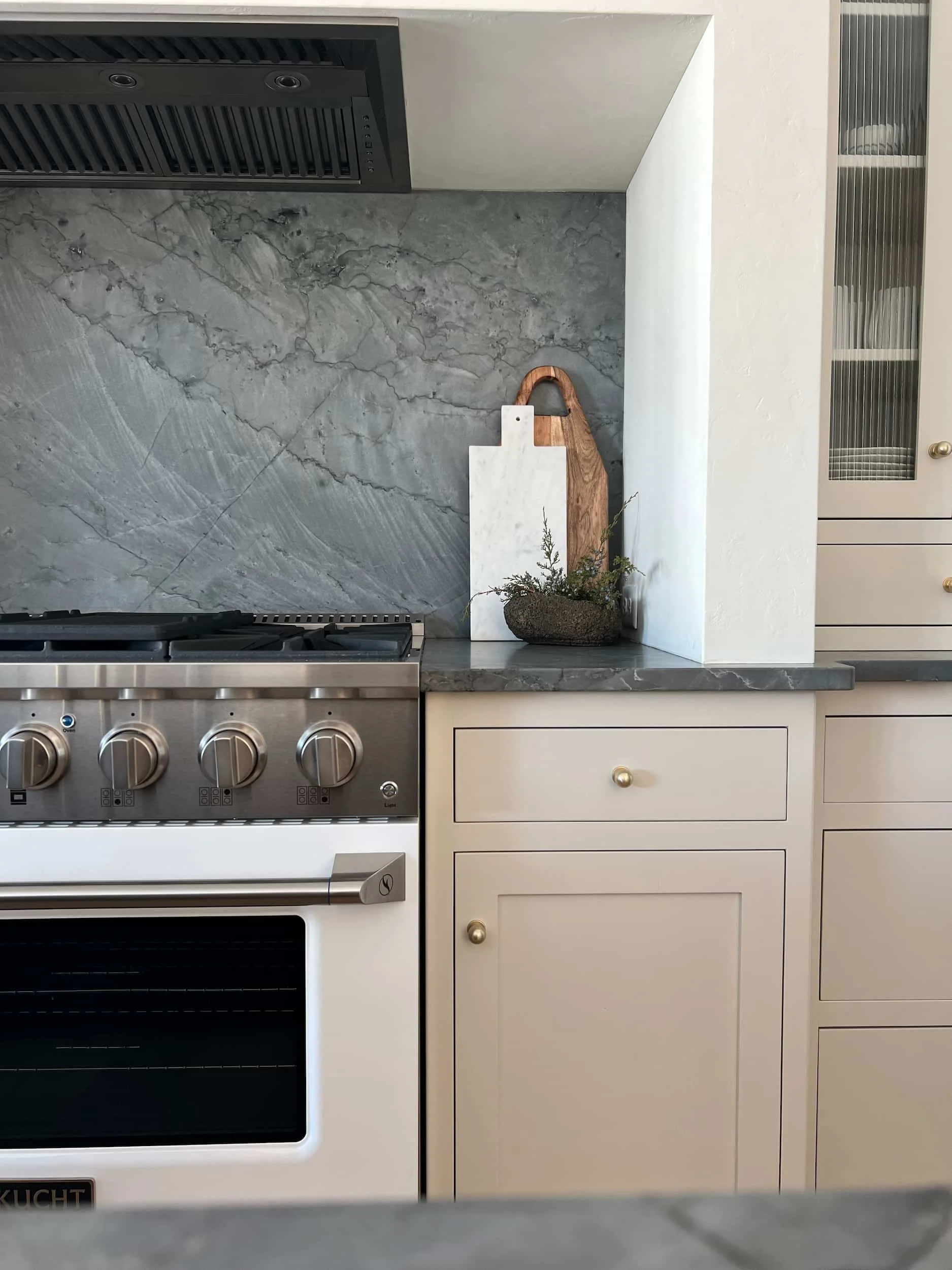Kitchen range area featuring a large, dark gray soapstone slab backsplash and counter. The range is accented by light beige or white cabinetry and a white cutting board.