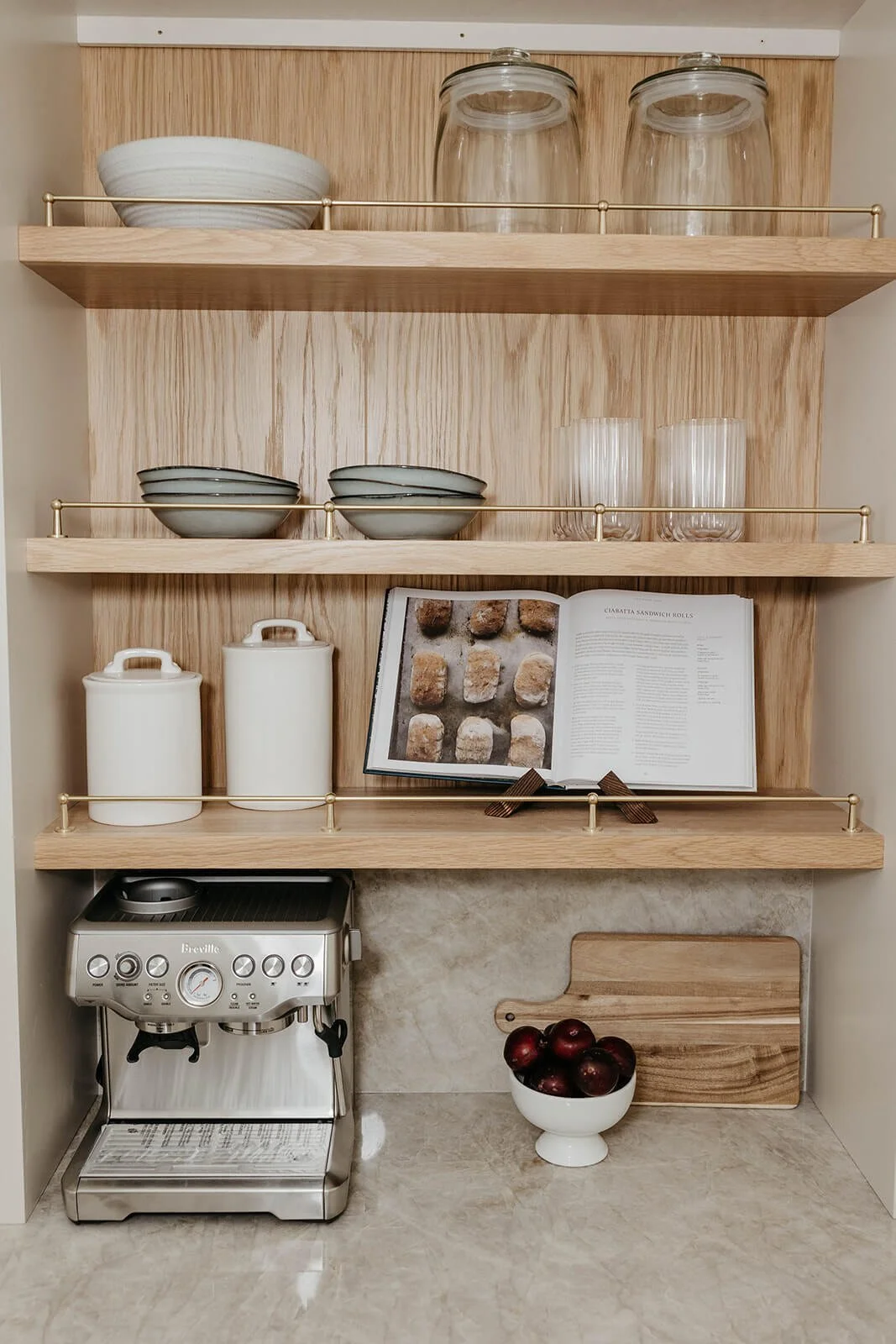 Custom coffee bar station with light wood veneered shelving featuring brass rails, a stainless steel espresso machine, and cookbooks. Dishes and glassware are stored above."