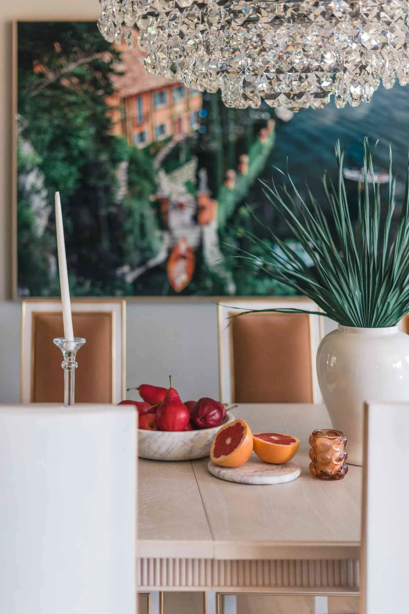 Styled dining table close-up featuring a marble bowl of red fruit, sliced grapefruit, a white ceramic vase with greenery, and a tall crystal candle holder. A large, crystal cloud-shaped chandelier hangs overhead.