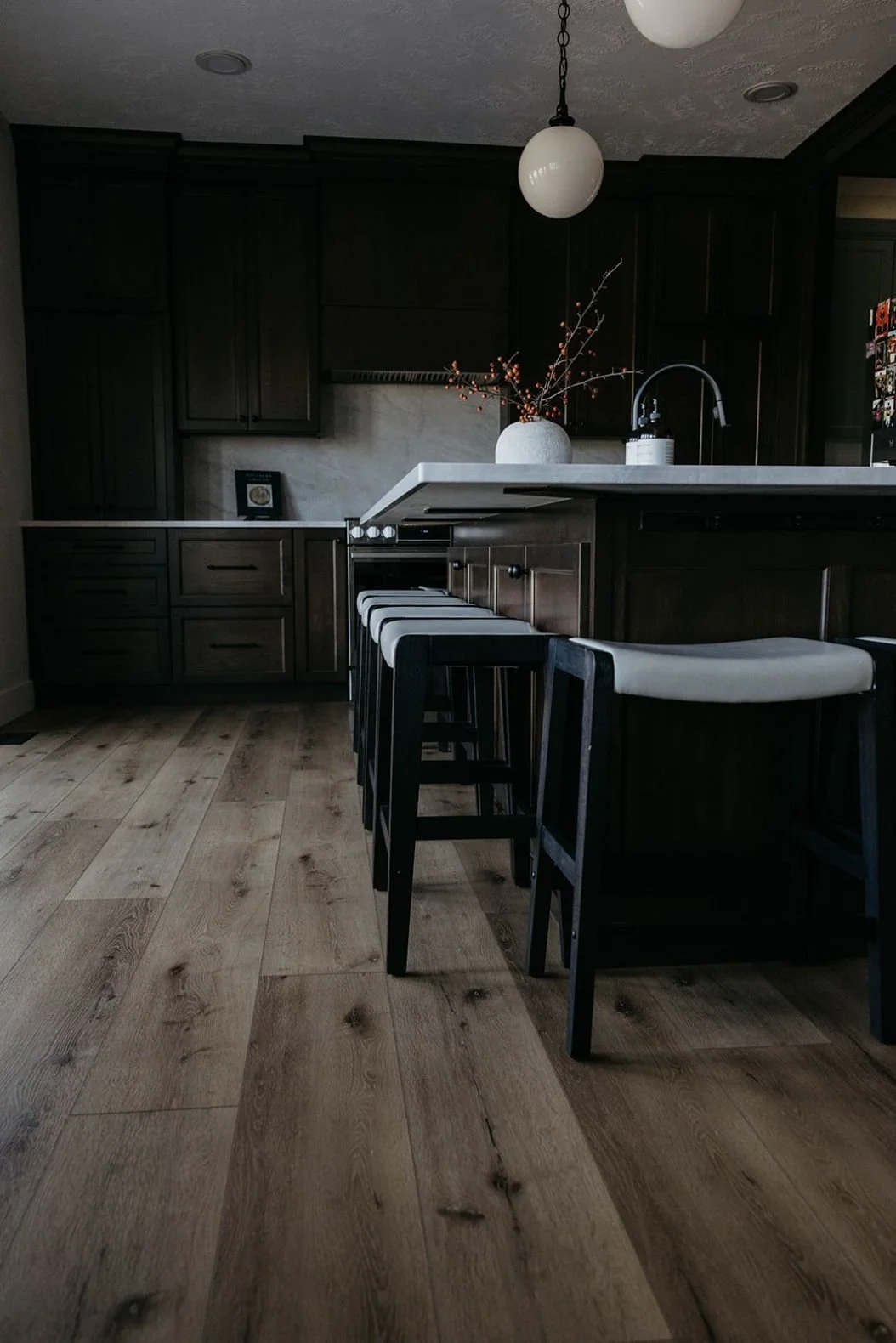 View of the kitchen island with three counter stools and a modern white globe pendant light hanging overhead. The counter is styled with a vase of branches.