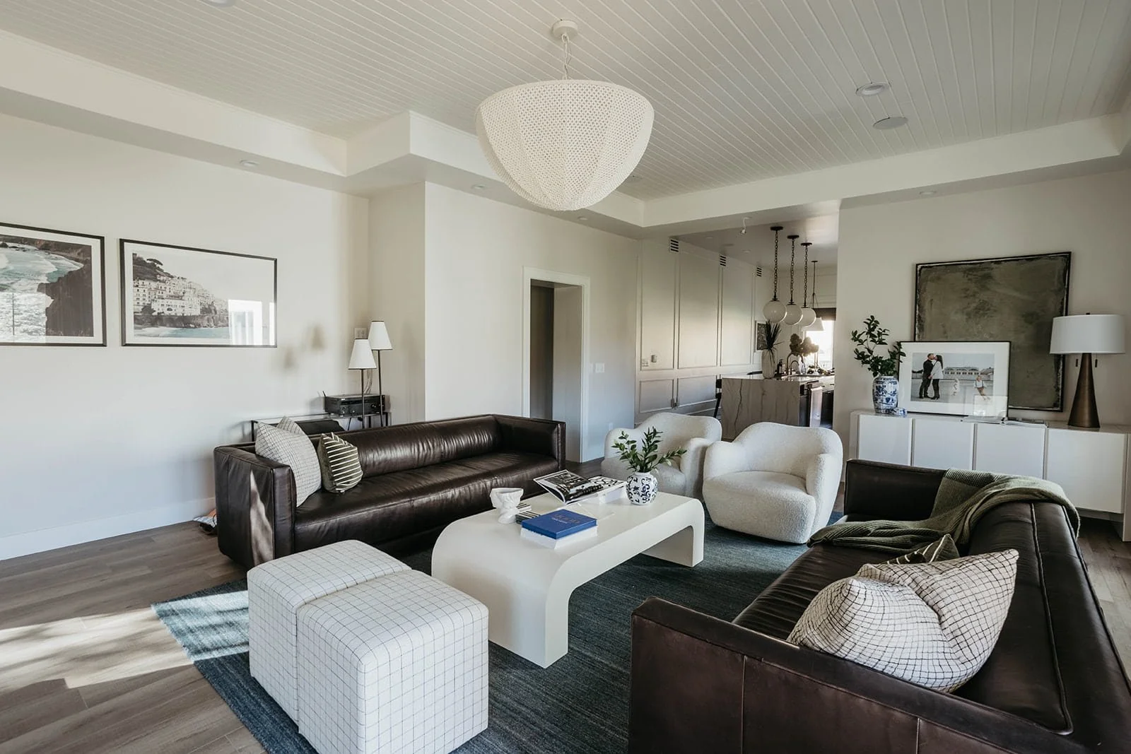 Bright, open-concept living room featuring a dark leather sofa, white accent chairs, a coffered ceiling, and a unique cloud-shaped white chandelier.