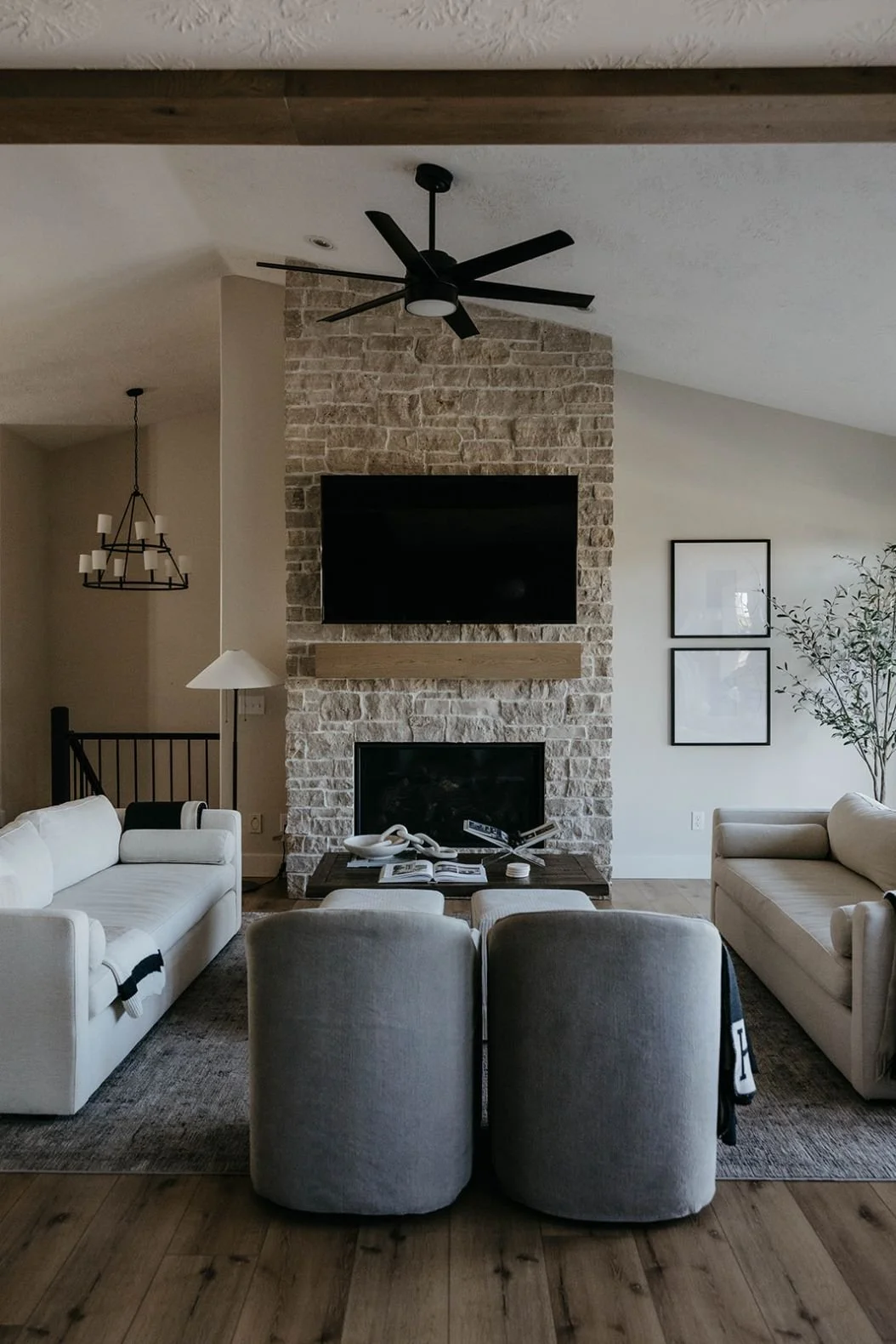 Living room with a stone-clad fireplace as the focal point, flanked by large windows. The area includes a white sofa and two round gray swivel chairs.