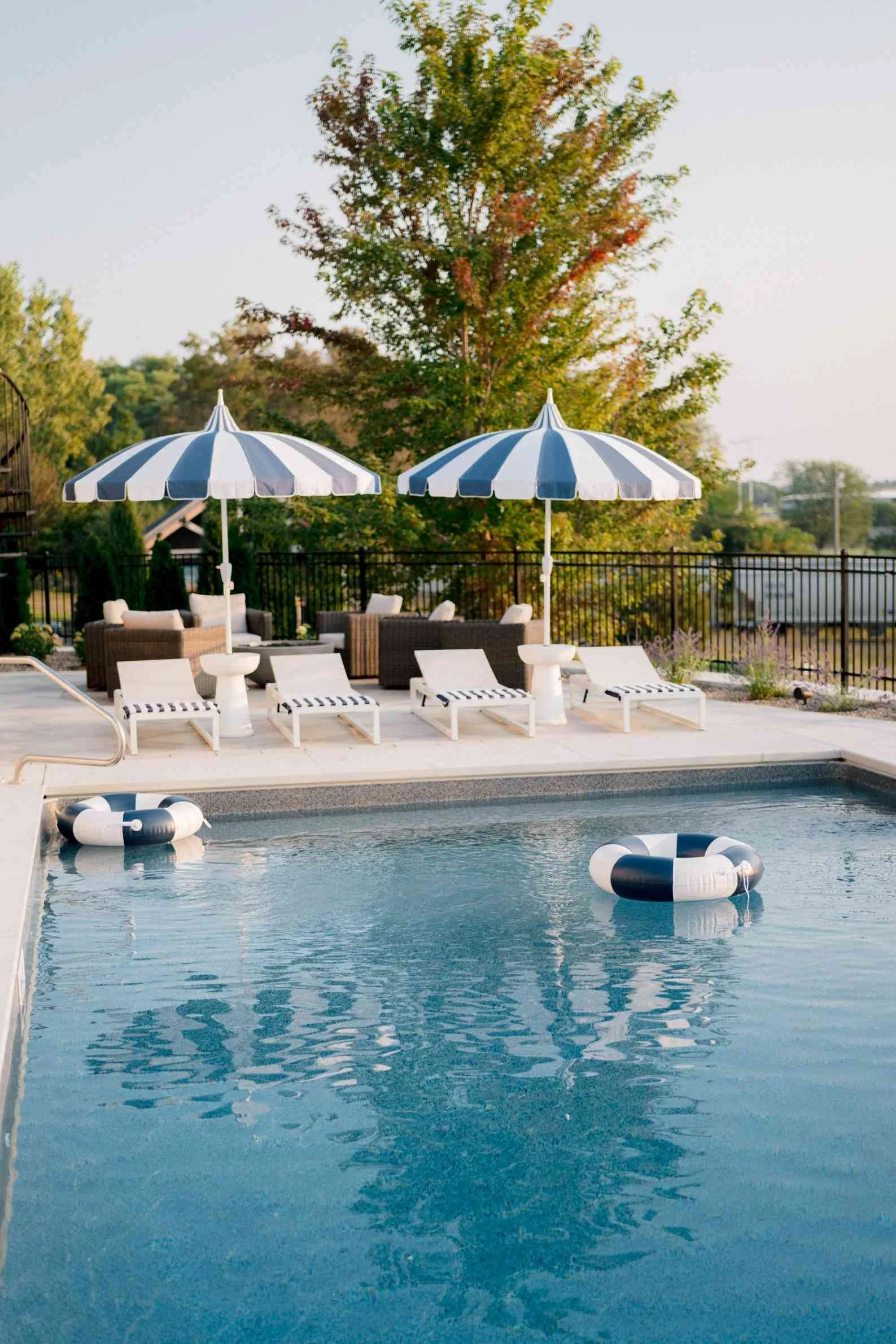 Stylish pool lounge area with two navy and white striped umbrellas shading white chaise lounges. Two striped ring floats are visible in the pool.