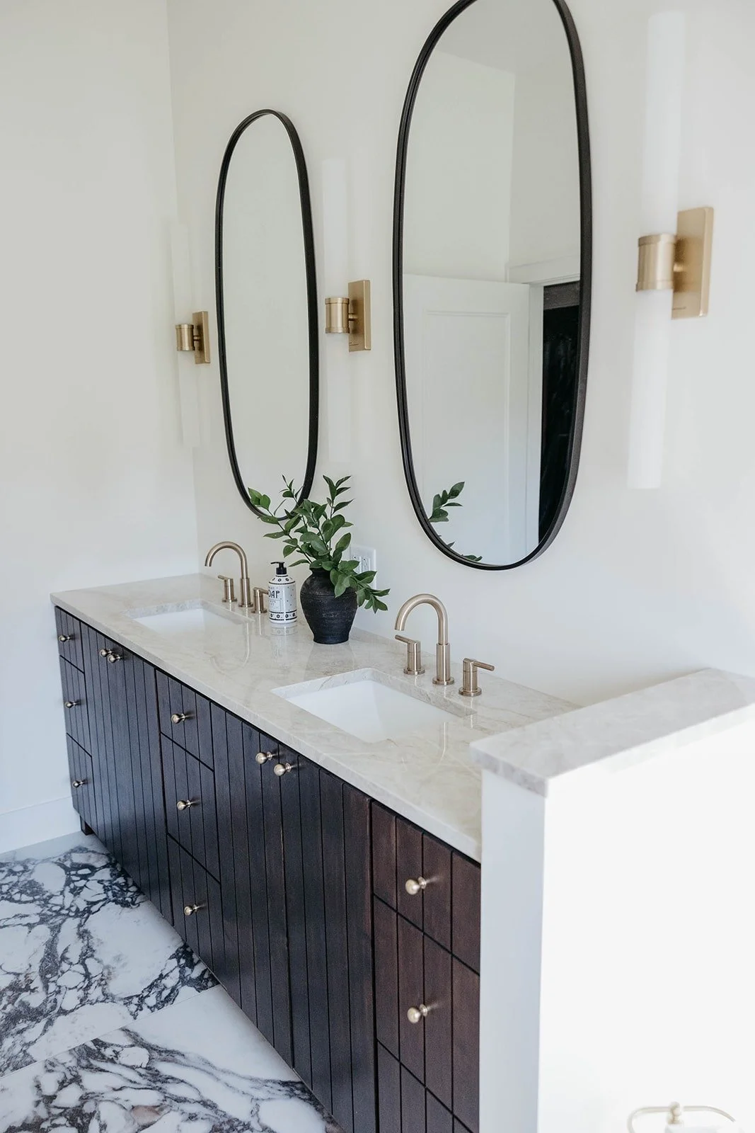 Wide view of a modern bathroom double vanity with dark wood cabinetry and a white marble countertop. Two tall, oval black-framed mirrors are flanked by modern brass sconces.