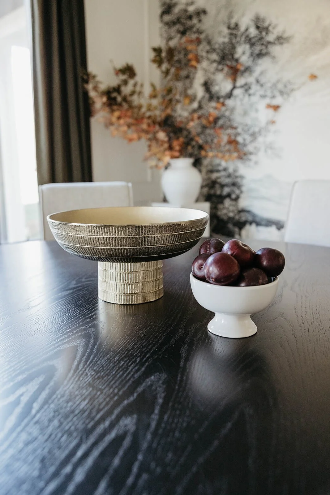 Close-up of a dark dining table styled with a textured gold pedestal bowl and a white bowl of red fruit. A floral arrangement and the nature mural are visible in the background.