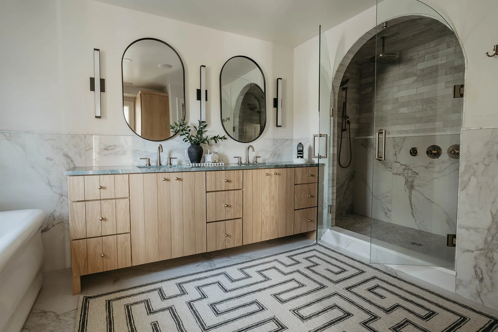 Wide view of the master bathroom showing the natural wood vanity, patterned floor, and a walk-in shower with an arched marble entrance.