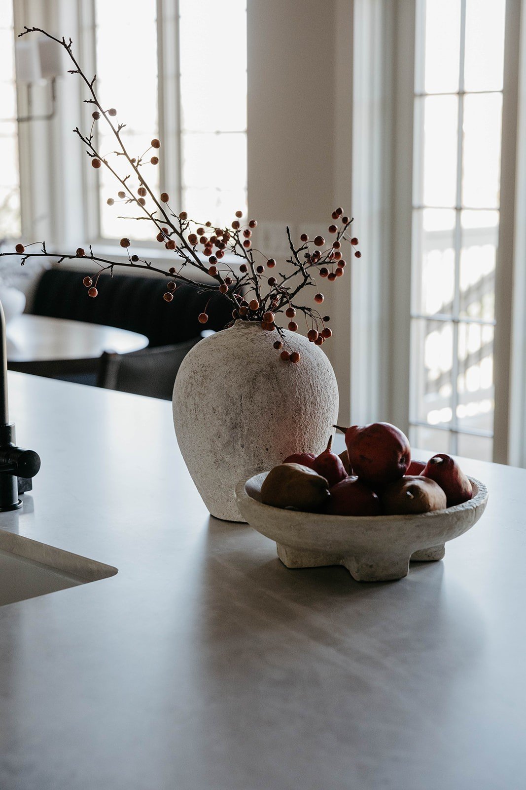 Close-up of kitchen countertop styling featuring a round stone bowl of apples and pomegranates next to a textured white vase with berries