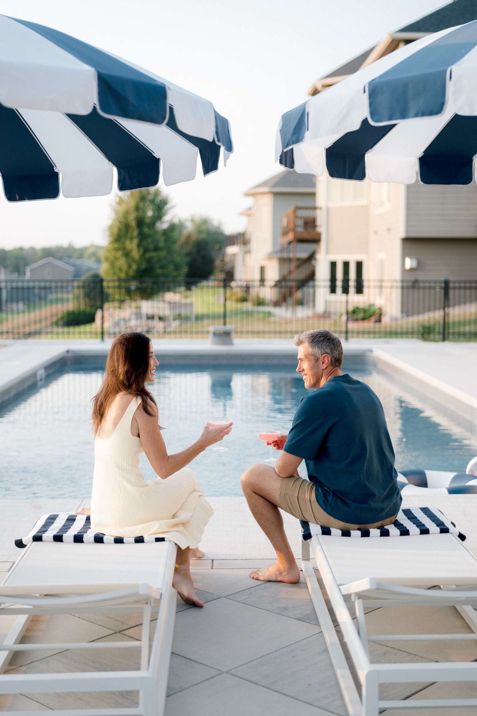 Christy Steen and husband sitting on white lounge chairs at the edge of the pool, talking and enjoying drinks on a sunny evening.