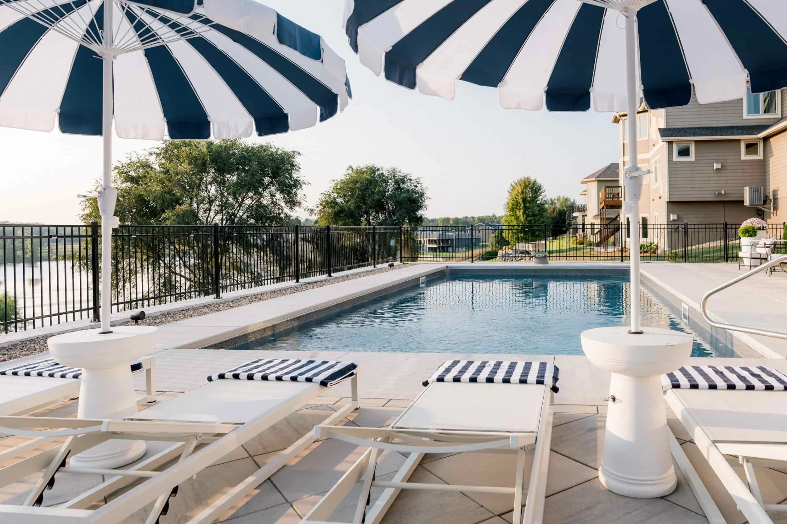 Luxury poolside scene with white lounge chairs, side tables, and navy and white striped umbrellas next to a rectangular swimming pool.