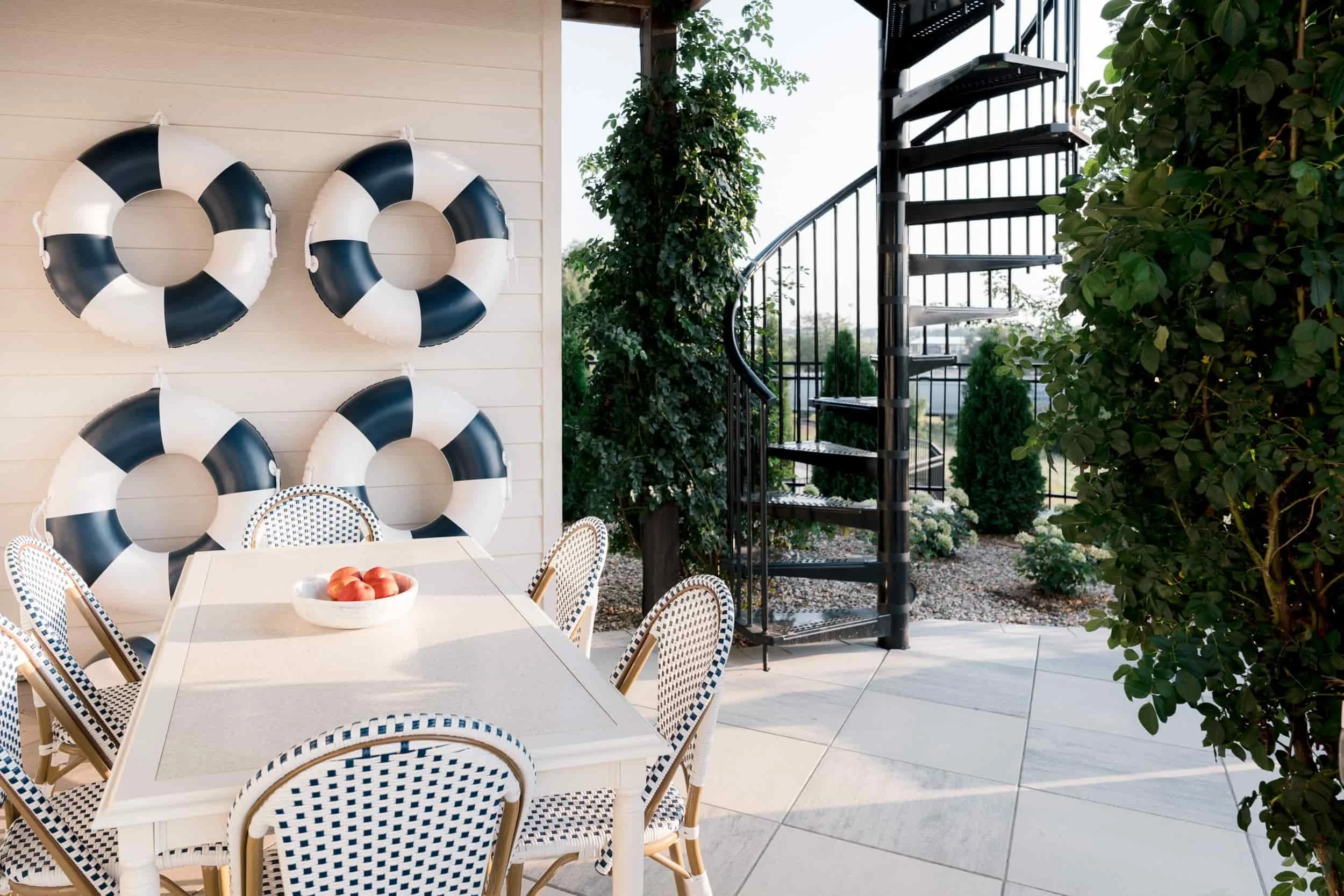Outdoor patio with a white dining table, navy and white woven chairs, and a whimsical wall of life preserver decor. A black metal spiral staircase is visible on the right, surrounded by lush greenery.