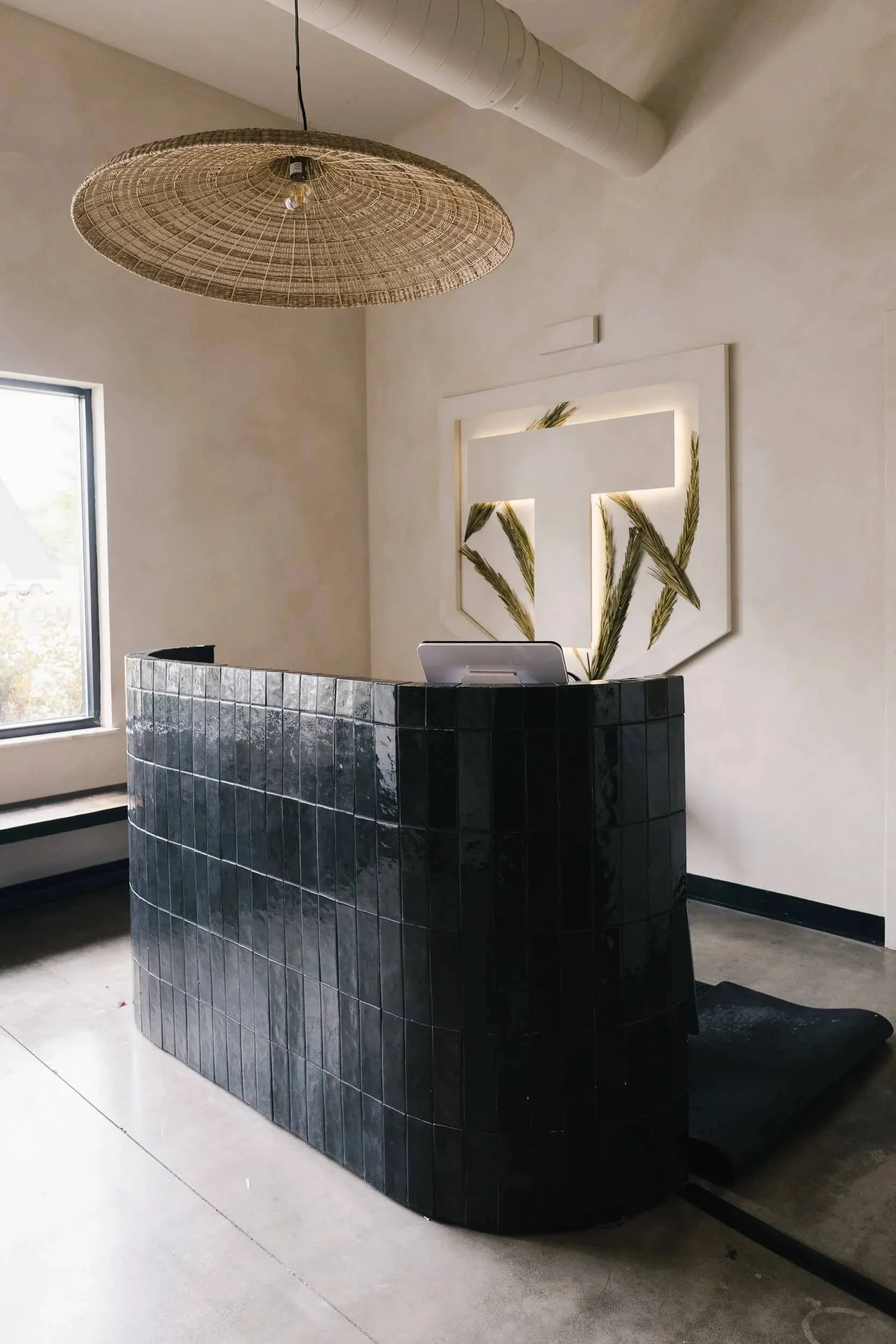 Modern reception desk tiled in glossy black subway tiles with a curved front, set against a light textured wall with a rattan chandelier overhead.