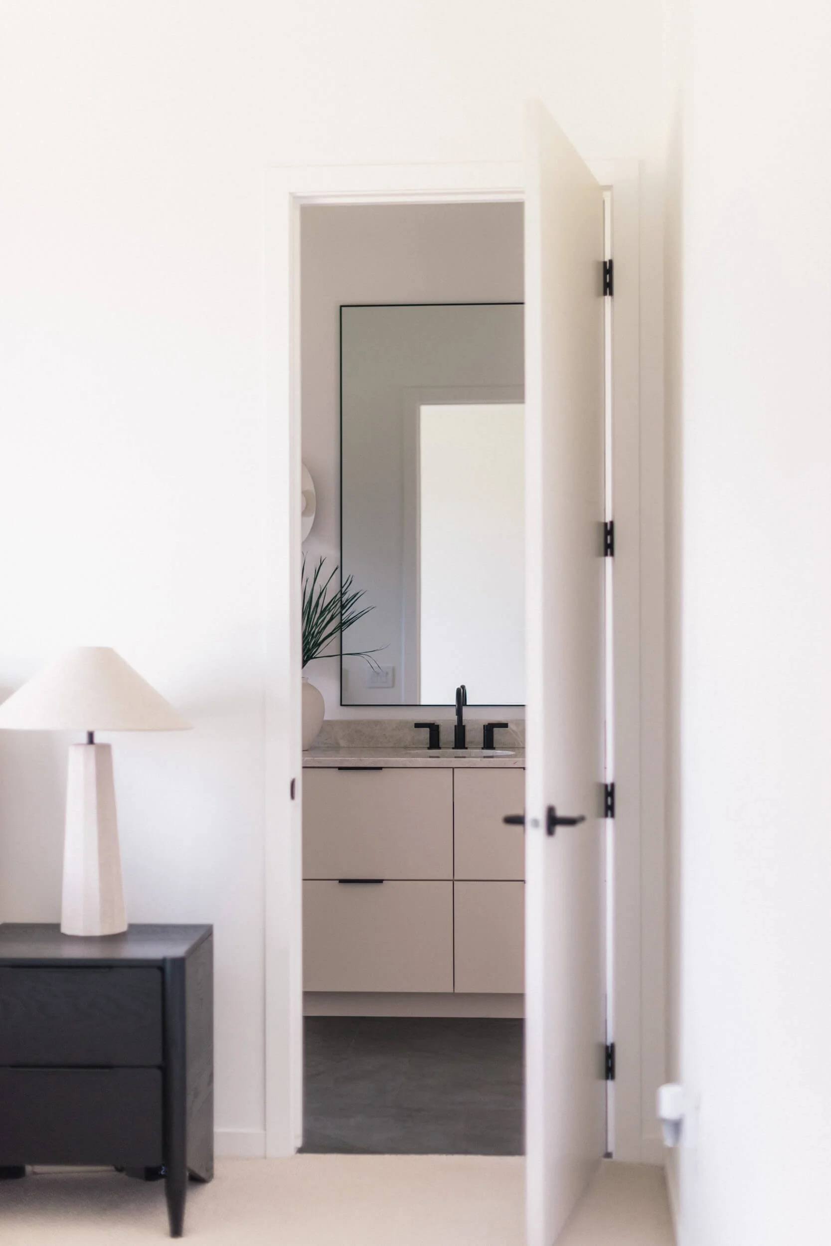 View into a modern bathroom with light taupe double vanity and a mirror, seen past a black nightstand and white table lamp in the foreground.
