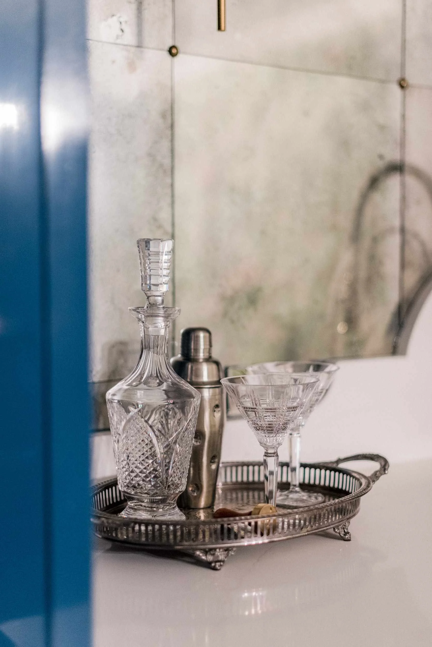 Close-up of a silver tray on the bar holding a crystal cocktail shaker, decanter, and coupe glasses, reflected in the antique mirror.