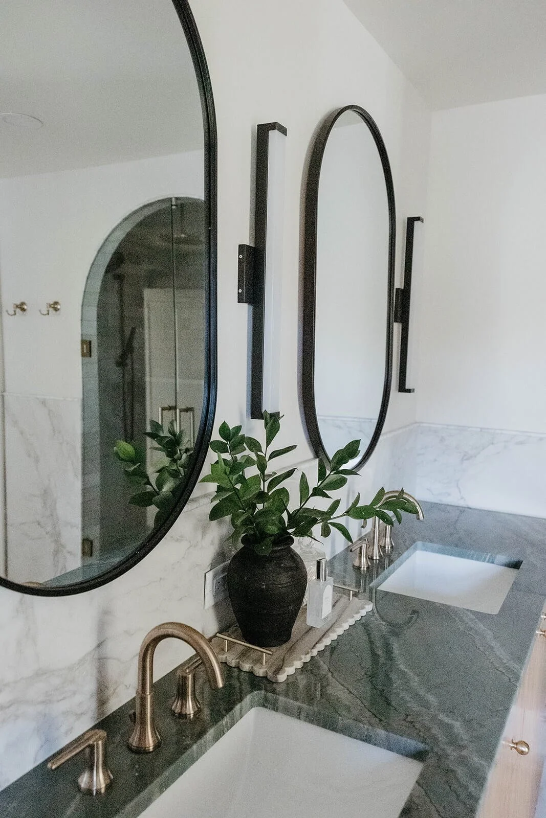 Double vanity bathroom featuring a green marble countertop, under-mount sinks, and a mix of black arched and oval mirrors hanging on a white marble wainscoting wall.