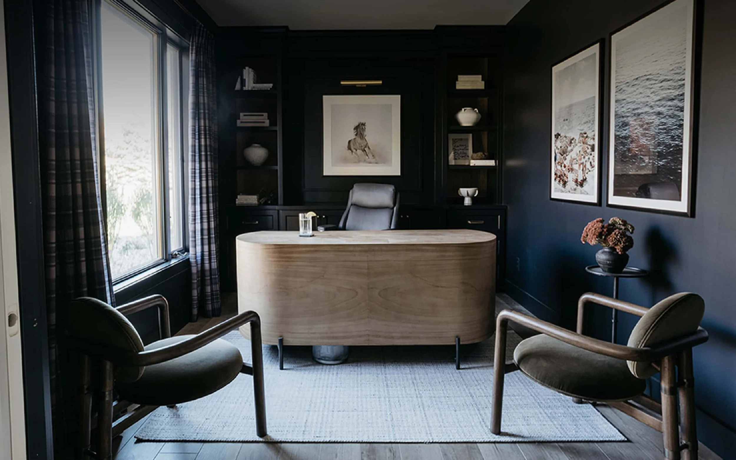 Moody home office featuring dark charcoal walls and built-in shelving, a light wood curved desk with a black base, and two mid-century modern style wood and velvet armchairs. The walls are decorated with large framed artwork, including two coastal pr