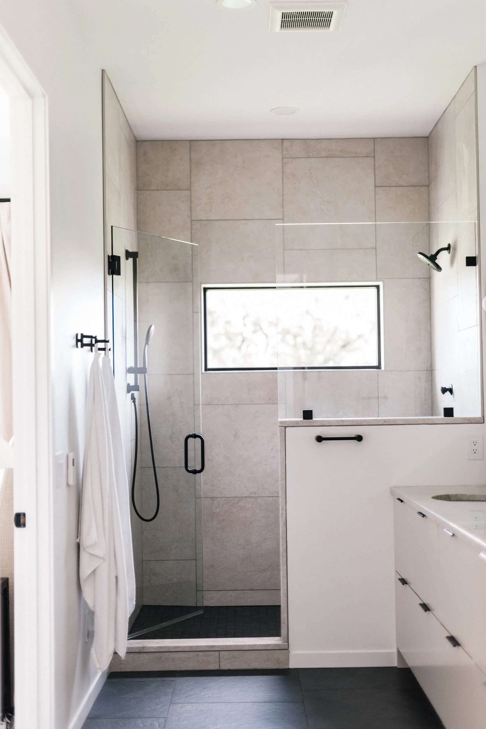 Walk-in shower area with light beige rectangular tiles, a thin rectangular window, modern shower floor with small black tiles, a dark baseboard, and a clear glass shower door.
