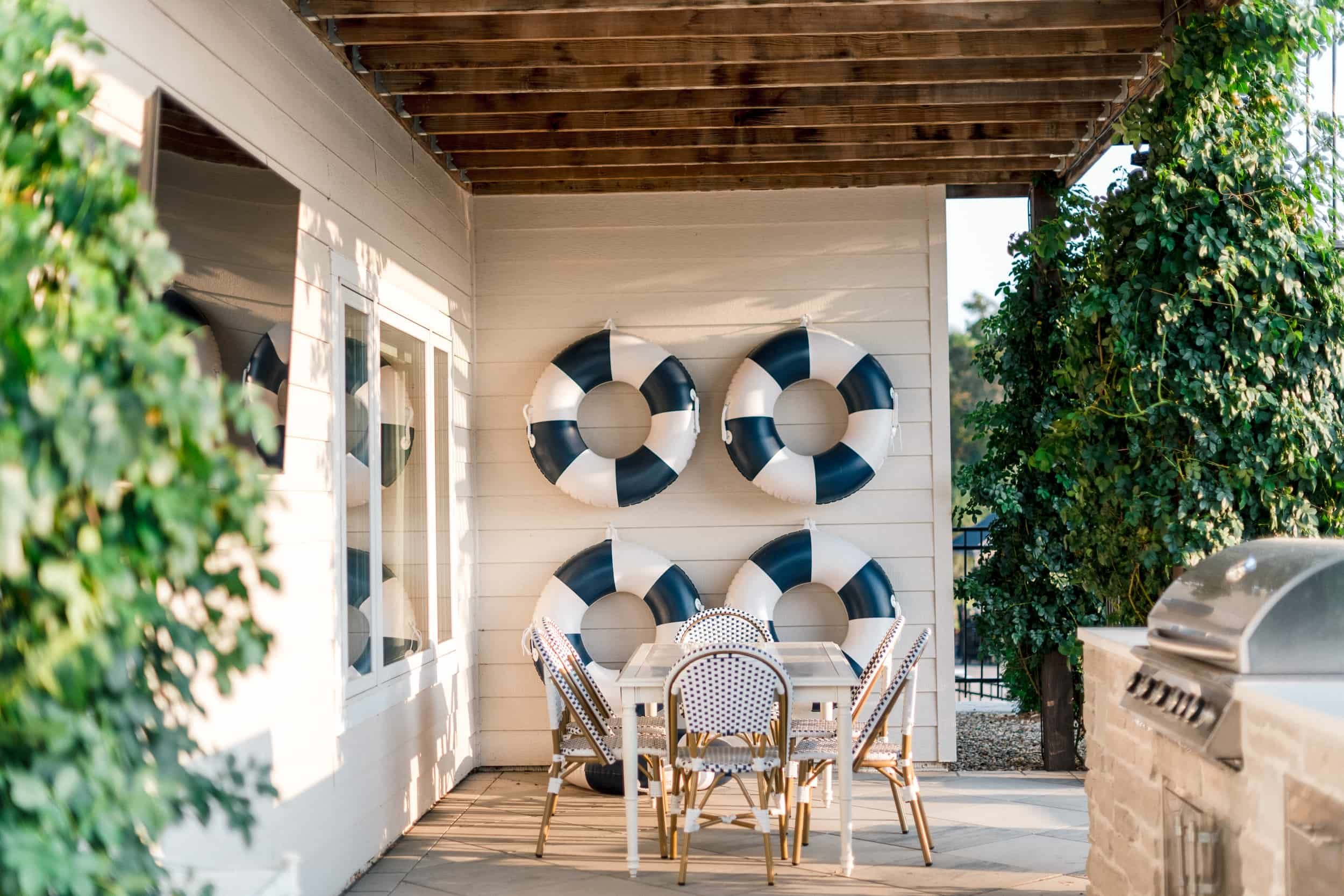 Covered outdoor patio dining area featuring a white shiplap wall decorated with a cluster of navy and white life preservers. The space includes a light-colored dining table and woven bistro chairs.