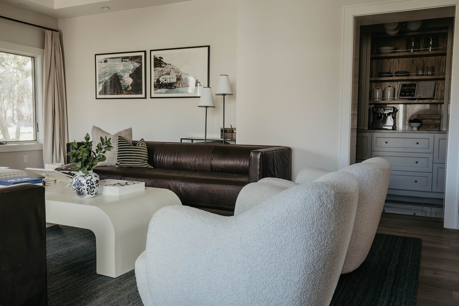 iew of the living room seating area featuring the dark leather sofa, two white curved bouclé chairs, and a white, sculpted coffee table.