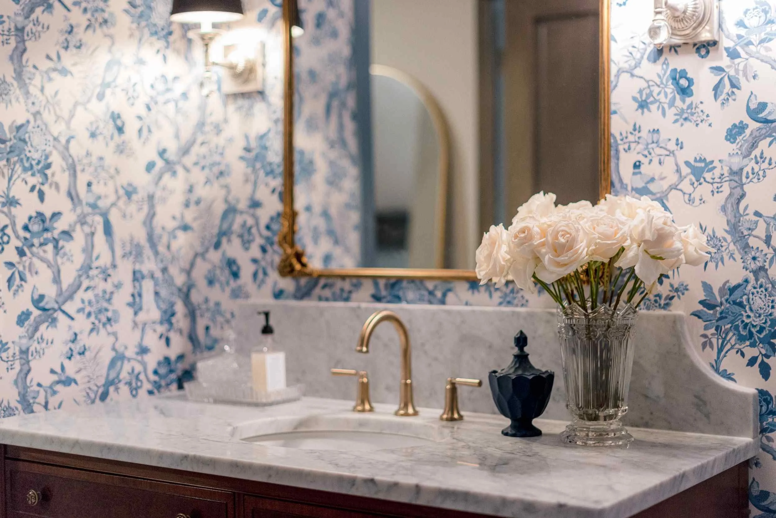 Detail of the white marble countertop, highlighting the crystal vase of cream roses, brass faucet, and navy blue vanity accessory.