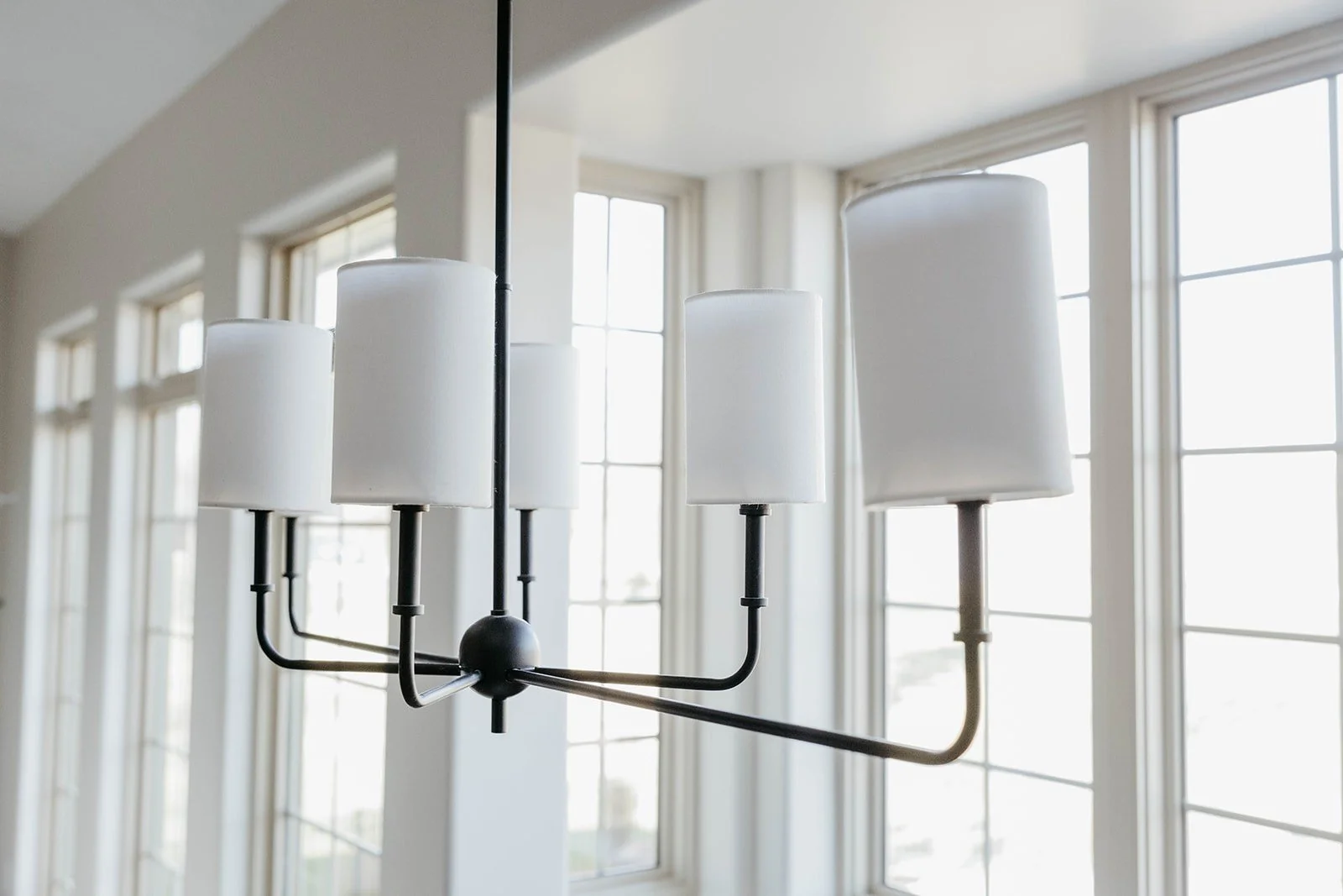 Close-up of a modern black linear chandelier with six white cylindrical shades, visible against large windows.