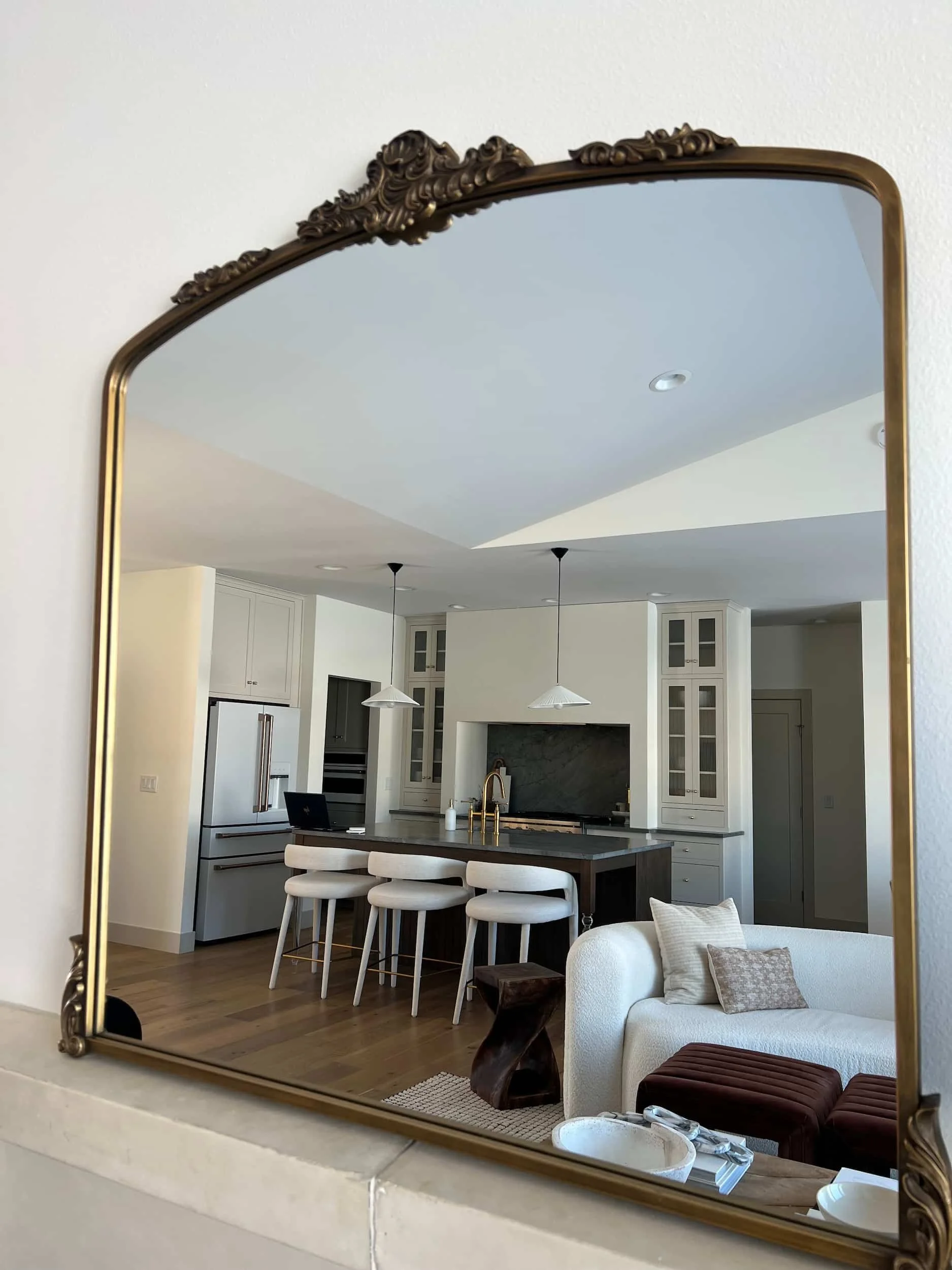Large ornate gold-framed mirror reflecting the bright, open-concept kitchen and living area, including the white cabinetry and dark island.