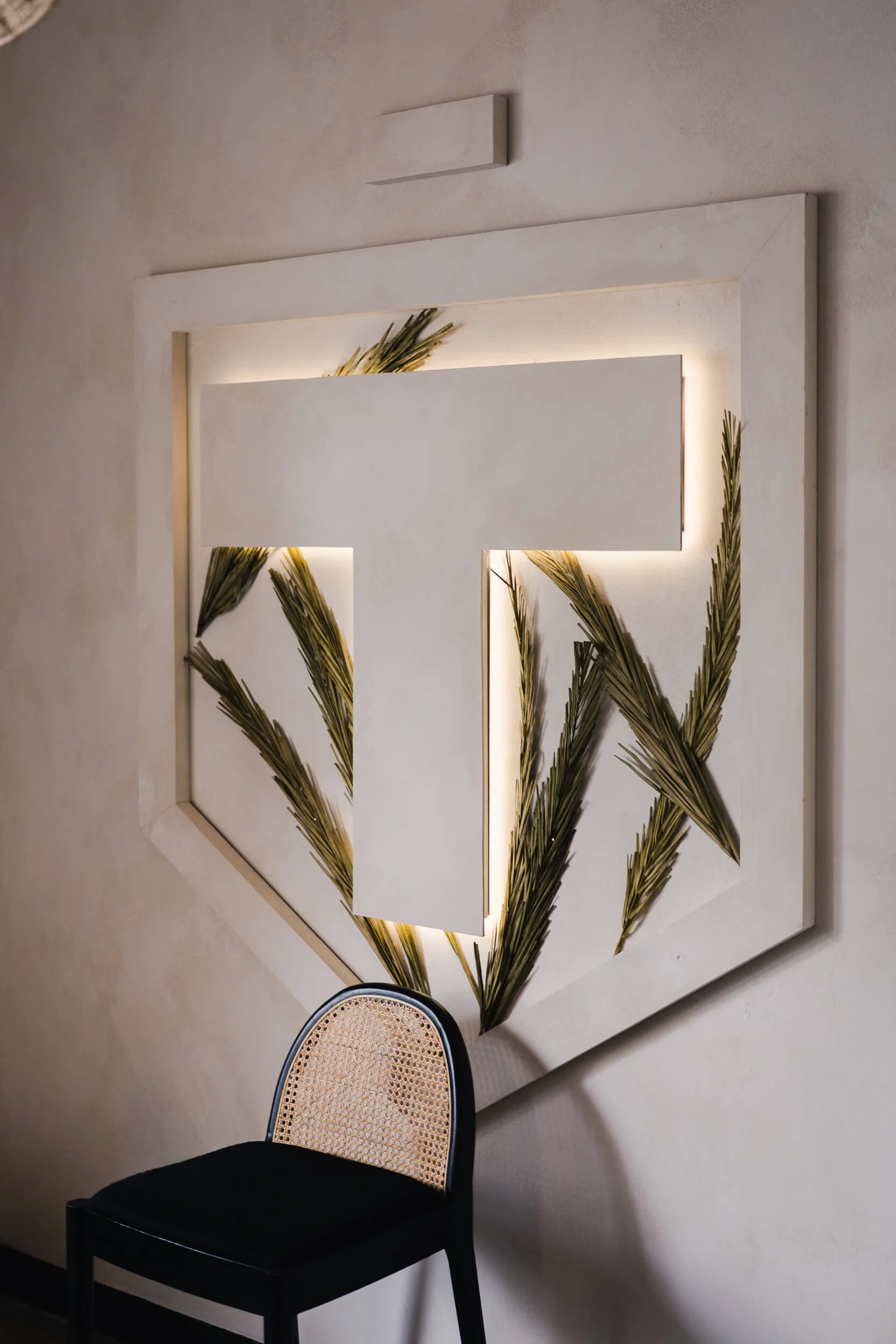 Close-up of the stylized 'T' logo in a white geometric frame with wheat stalks and backlighting, next to a black and cane chair.