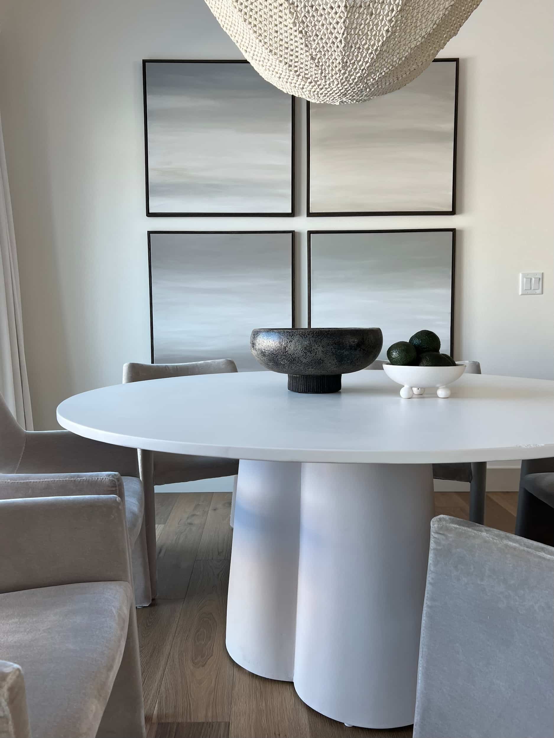 Close-up of the white round dining table with a sculptural base, styled with a dark textured bowl and decorative objects, set beneath a series of four framed abstract artworks.